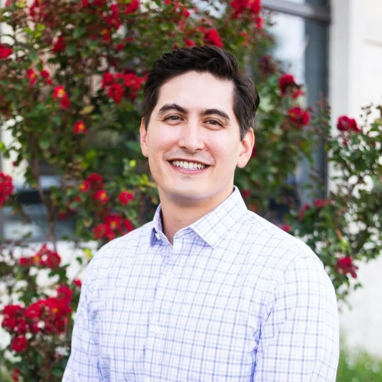 A young man with dark hair and a light checkered shirt smiling outdoors, with red flowering plants in the background.