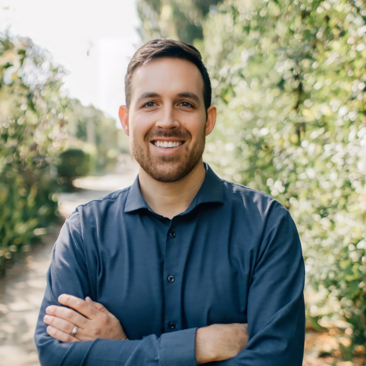 A smiling man with crossed arms standing outdoors in a lush, green environment.
