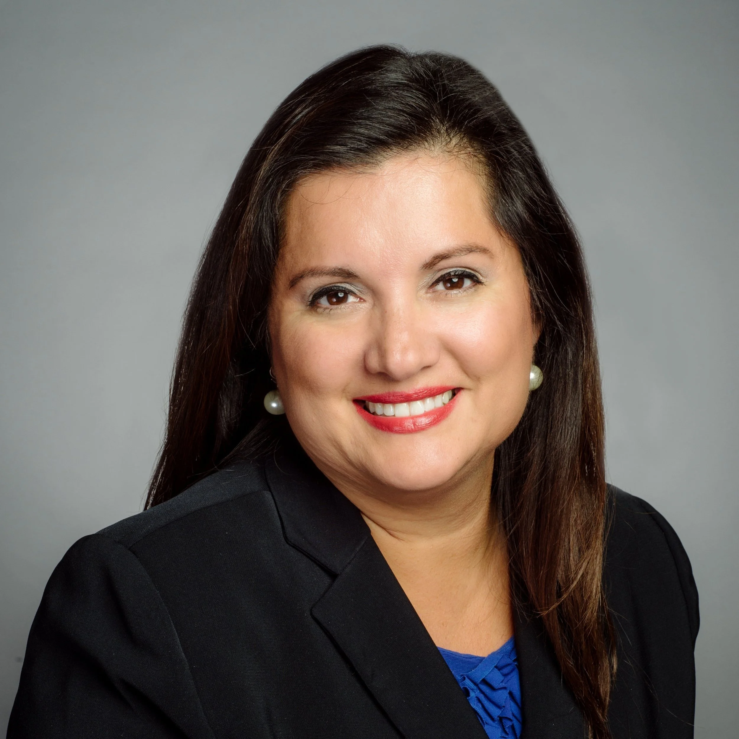 A woman with long dark hair wearing a black blazer, pearl earrings, and a blue top smiling at the camera against a gray background.