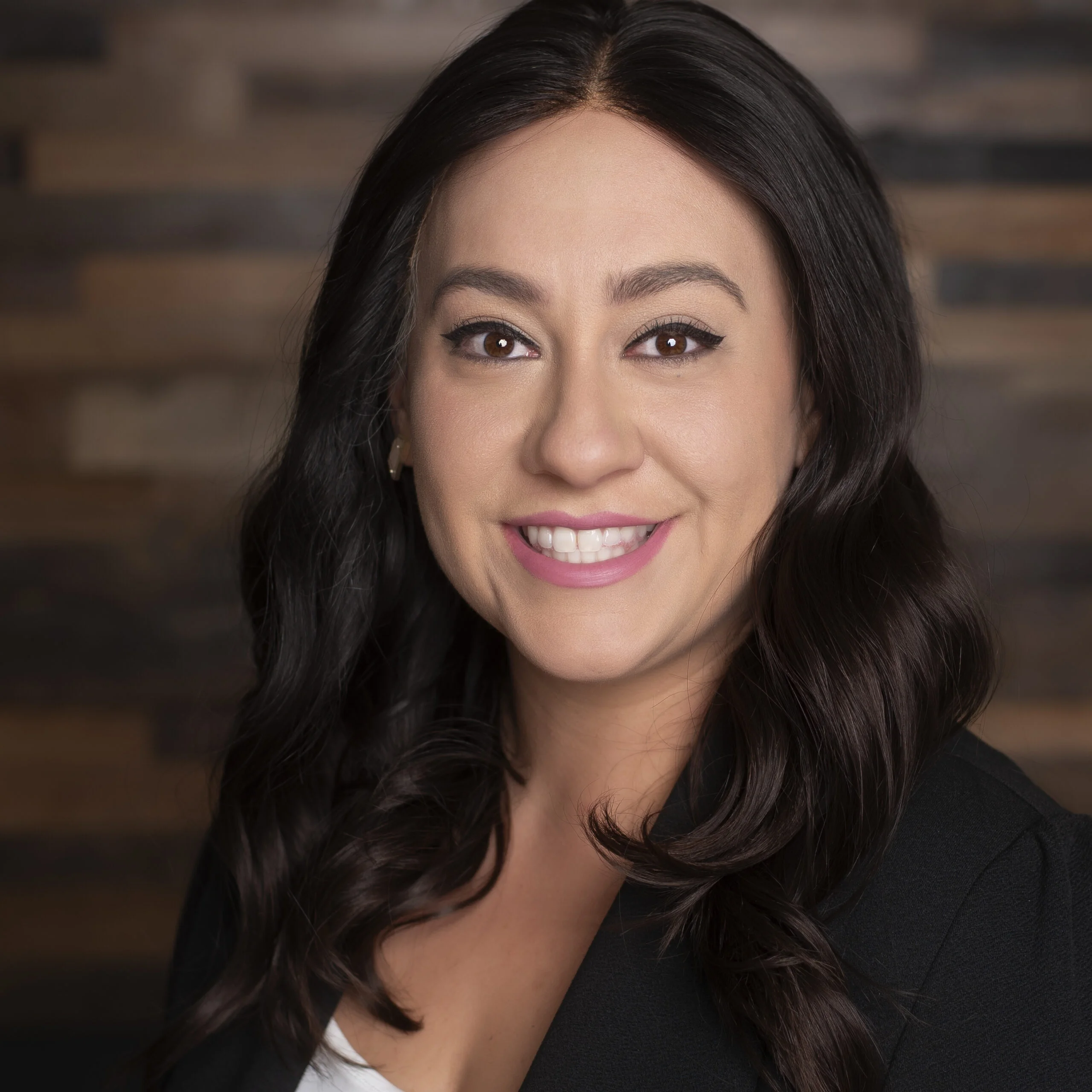 Close-up portrait of a smiling woman with dark brown, wavy hair, wearing a black blazer, against a blurred wooden background.