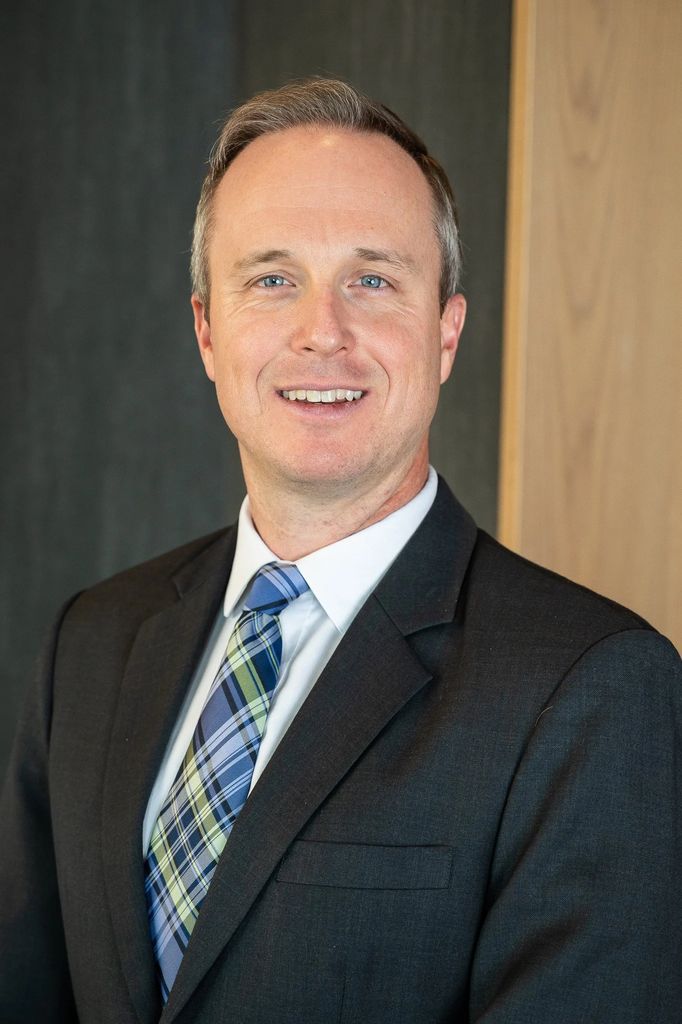 A man with short light brown hair and blue eyes, smiling, wearing a dark suit, white shirt, and a plaid tie, standing indoors against a background of dark and light wooden panels.