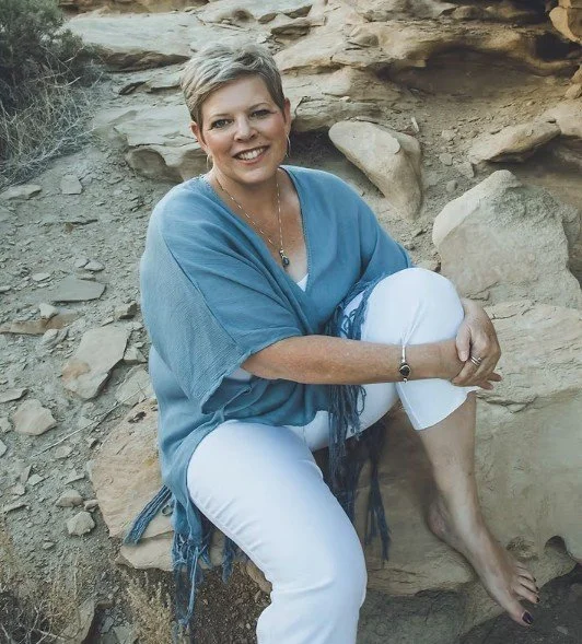 Woman sitting on rocks outdoors, smiling, wearing a blue top and white pants.