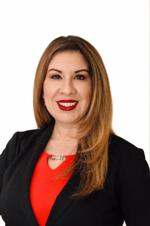 A woman with long, light brown hair, wearing a black blazer over a red top, smiling with red lipstick, against a white background.
