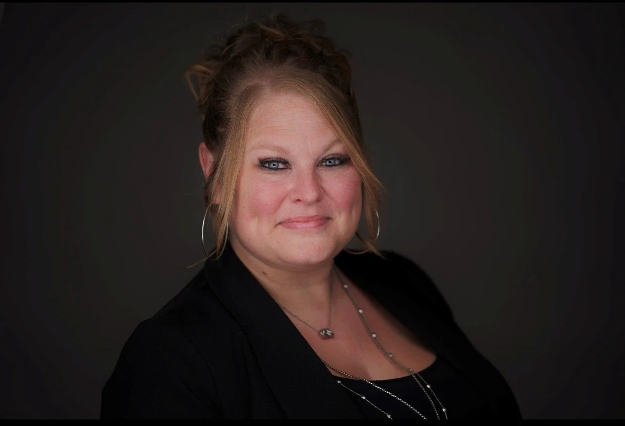 A woman with blonde hair styled in an updo, wearing hoop earrings, a black blazer, and layered necklaces, smiling at the camera against a dark background.