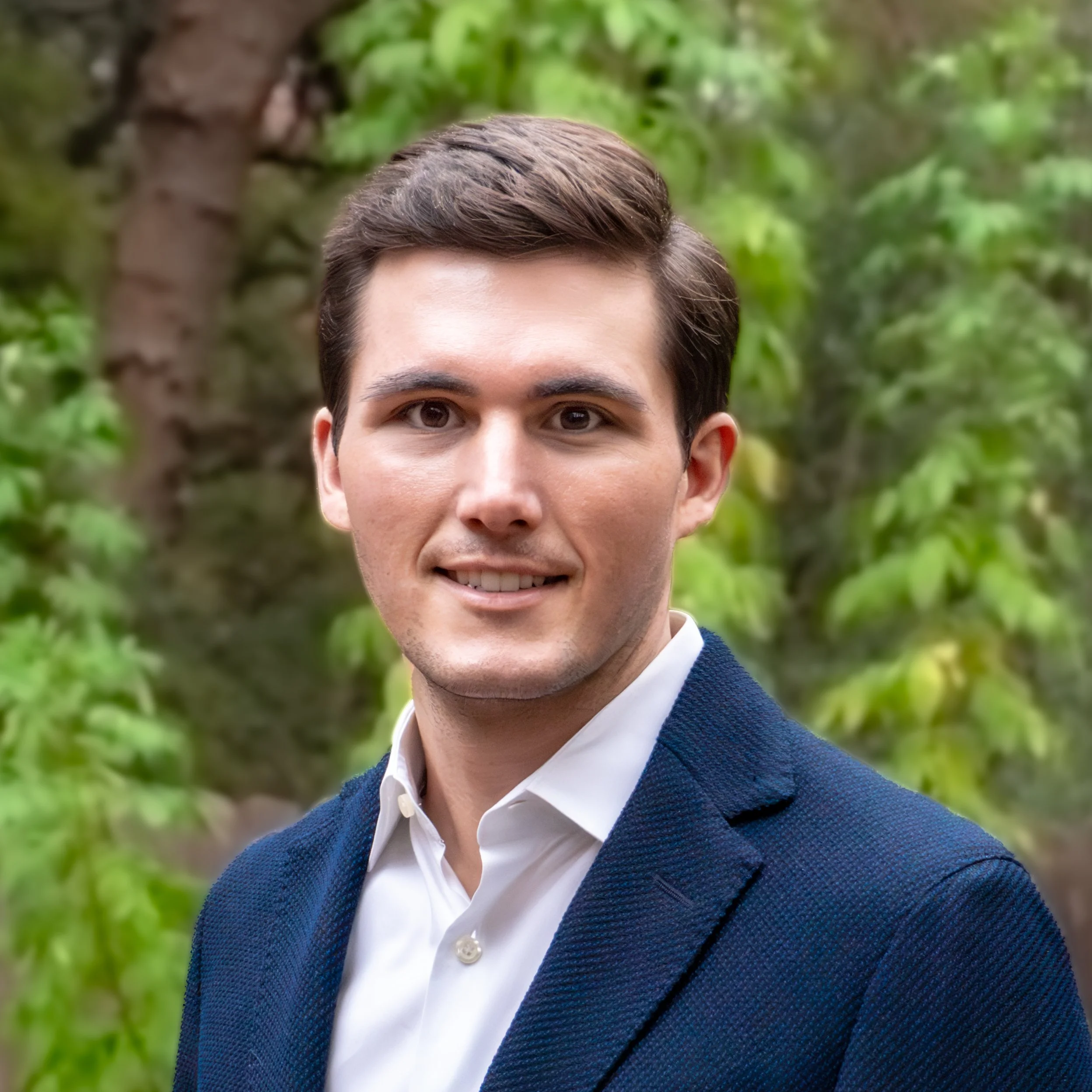 A young man with brown hair, wearing a white shirt and a dark blue blazer, is outdoors with green trees in the background.