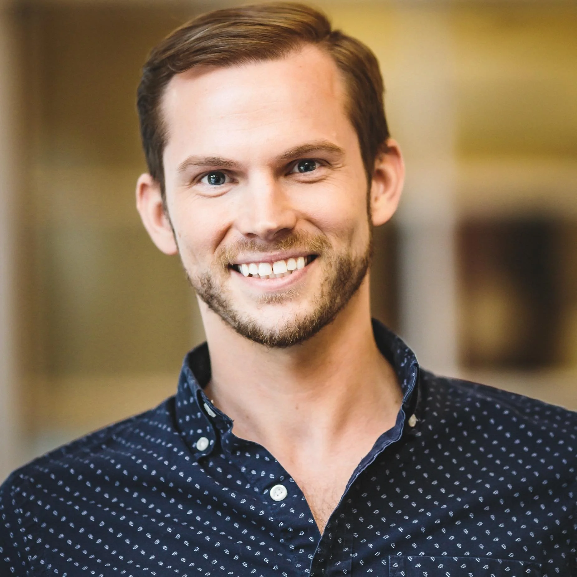 A smiling man with light brown hair and a beard, wearing a dark blue shirt with small white dots.