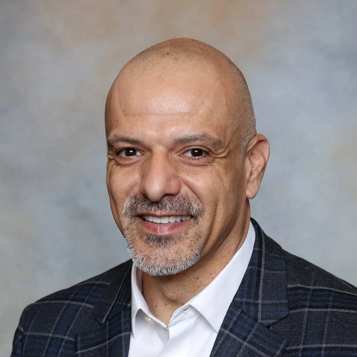 Close-up portrait of a middle-aged bald man with a goatee, smiling, wearing a dark checkered blazer and white shirt, against a neutral background.