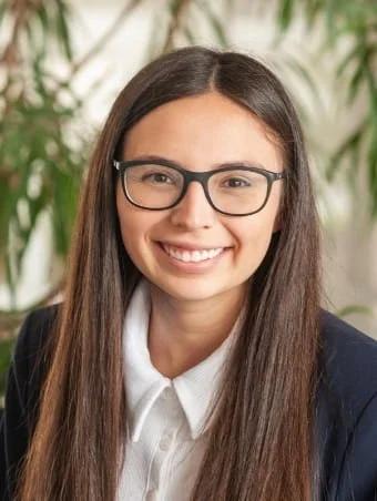 A young woman with long brown hair, glasses, and a bright smile, wearing a white collared shirt and a dark blazer, in front of green plants.