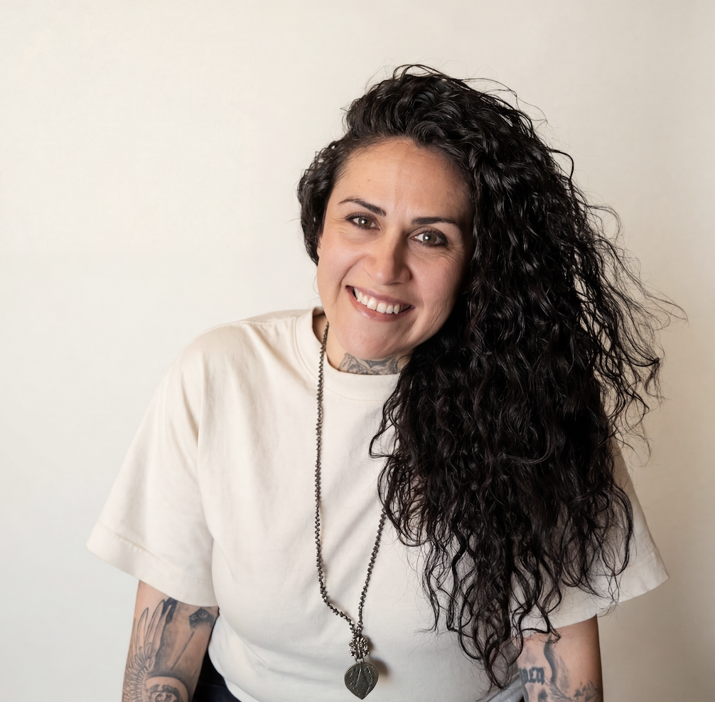 A woman with curly dark hair smiling, wearing a white t-shirt, tattoos on her arms, and a necklace with a leaf pendant, against a plain background.