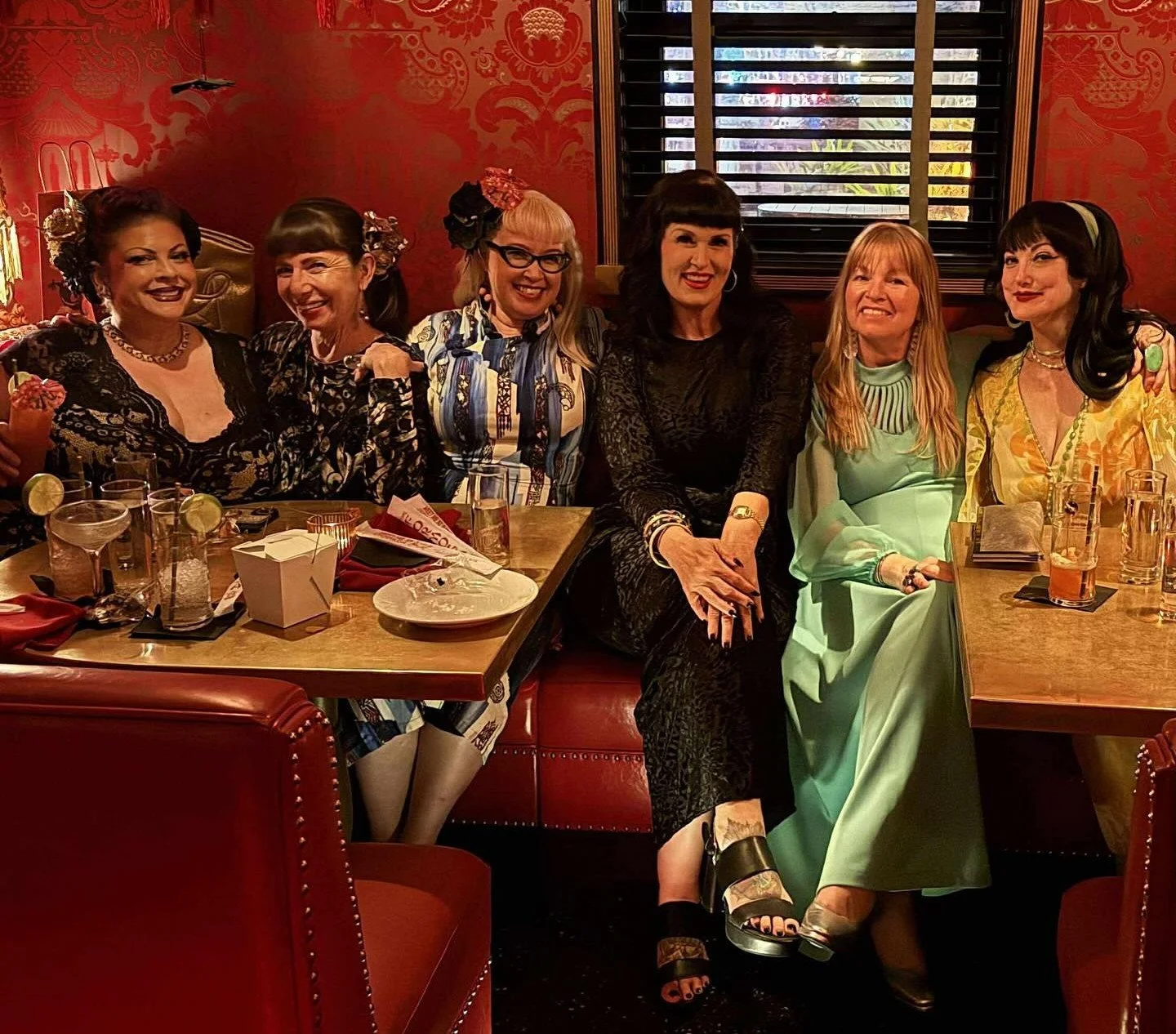 Six women in vintage attire smiling at a red leather booth inside The Formosa Cafe while enjoying their private dinner.