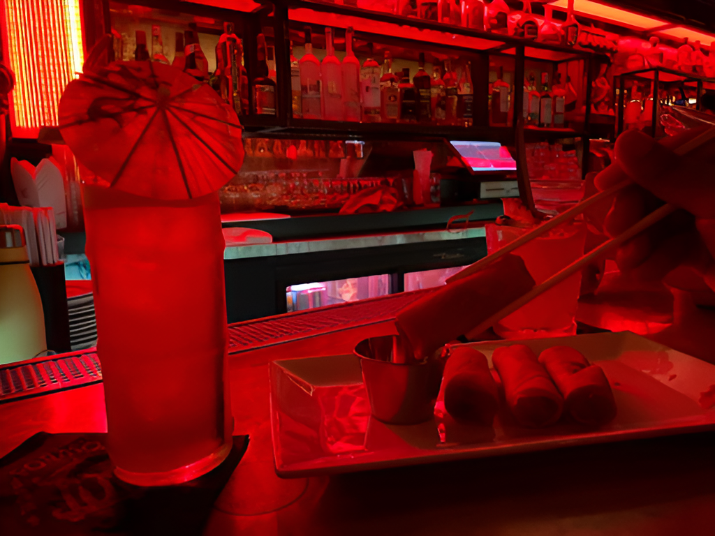 Red-lit bar with a tropical cocktail and chopsticks dipping an appetizer into sauce at The Formosa Cafe.