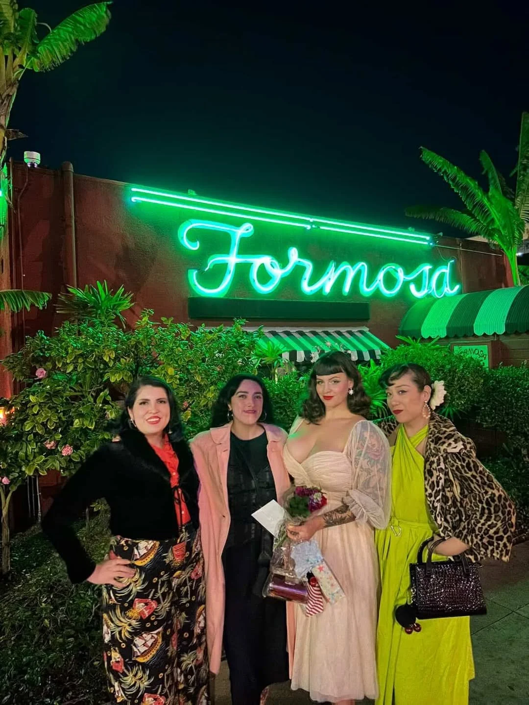 Four women posing beneath The Formosa Cafe’s glowing green neon sign on a lively West Hollywood evening.