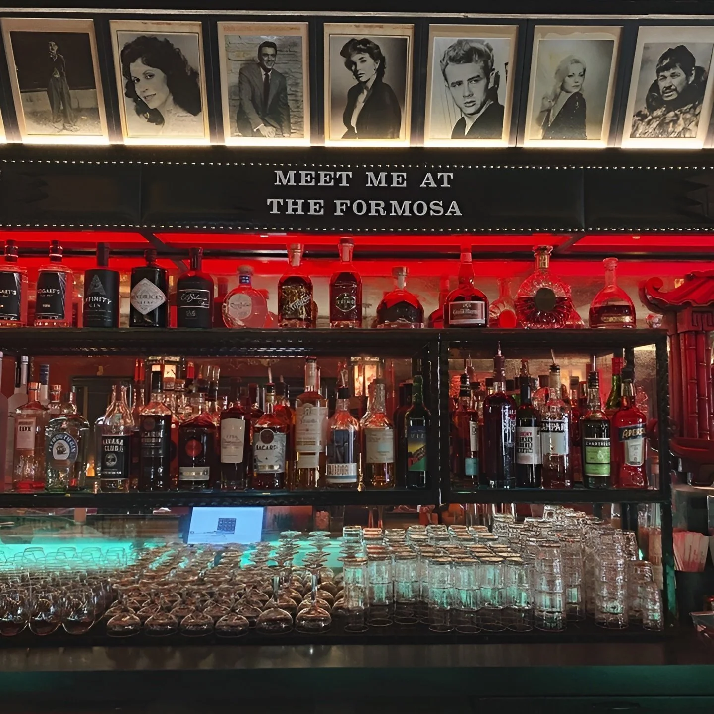 Red-lit bar shelves with liquor bottles and celebrity photos under a “MEET ME AT THE FORMOSA” sign at The Formosa Cafe.
