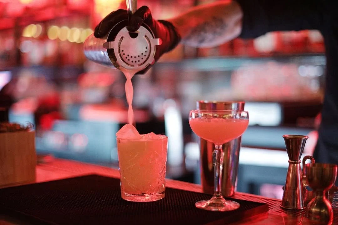 Bartender pouring a pink cocktail into a coupe glass at a moody, red-lit Hollywood lounge at The Formosa Cafe.