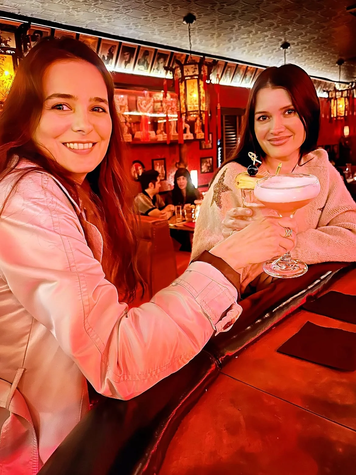 Two women smiling and enjoying cocktails at the red-lit bar counter inside The Formosa Cafe.