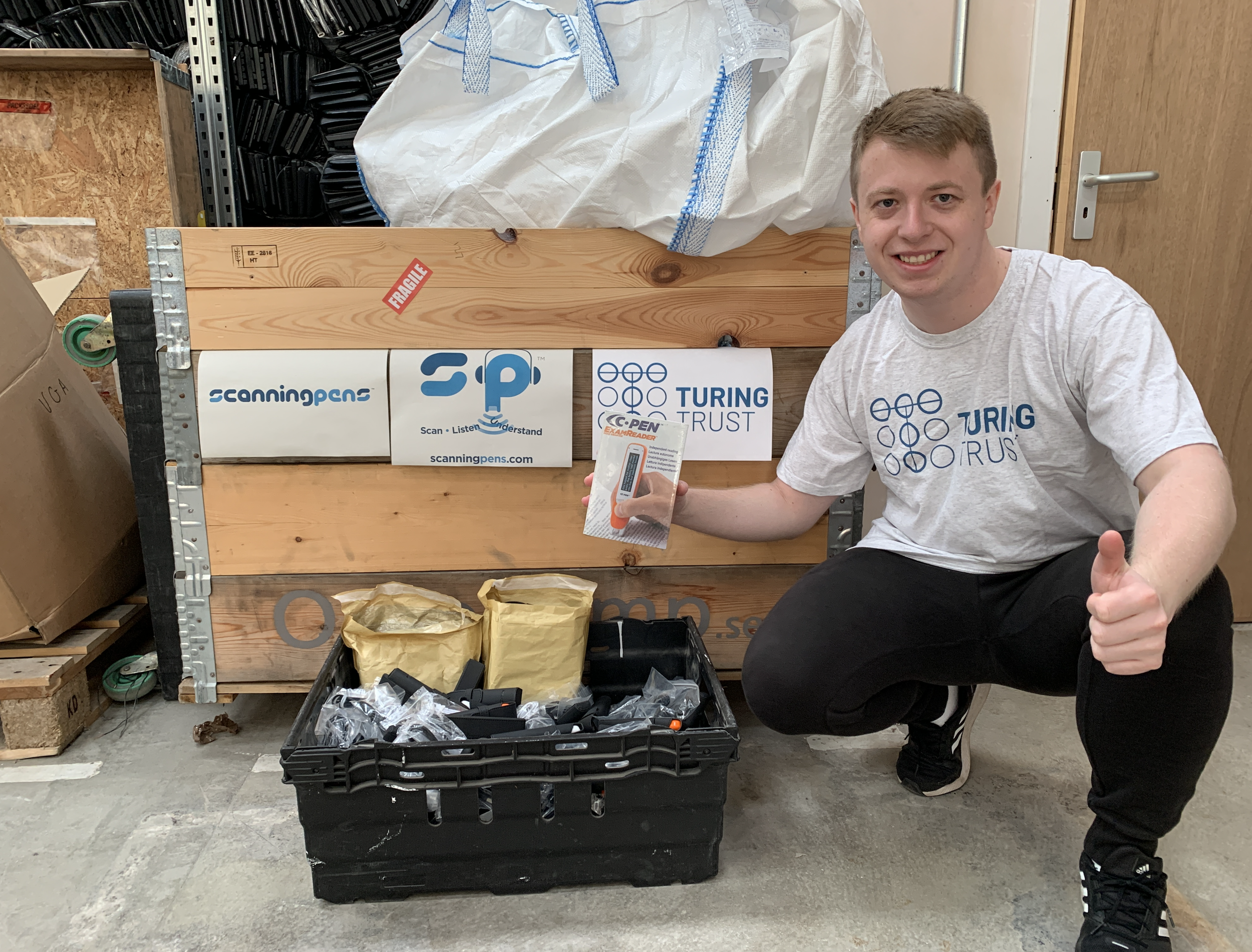 a person with short hair wearing a white T-shirt bearing the Turing Trust's logo kneeling down next to a crate of C-Pen Reader 2s and C-Pen Exam Reader 2s, giving a thumbs up and smiling at the camera