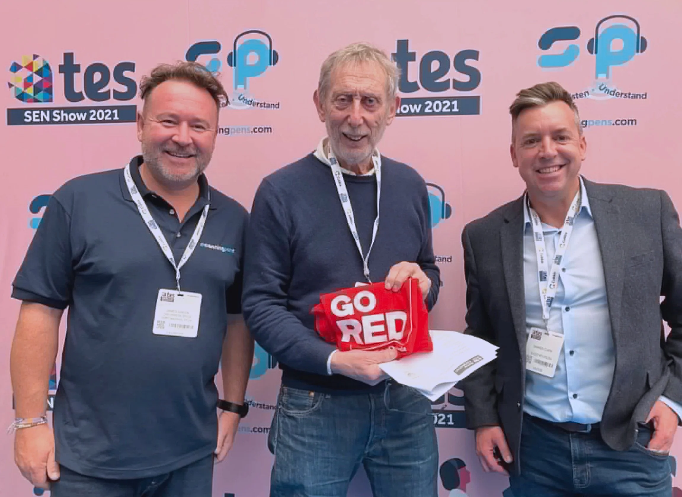 Three people dressed in casual clothes- James Green, Michael Rosen and Darren Clark- stood in front of a pink branded Scanning Pens and TES background. Michael is holding a Go Red for Dyslexia T-Shirt.