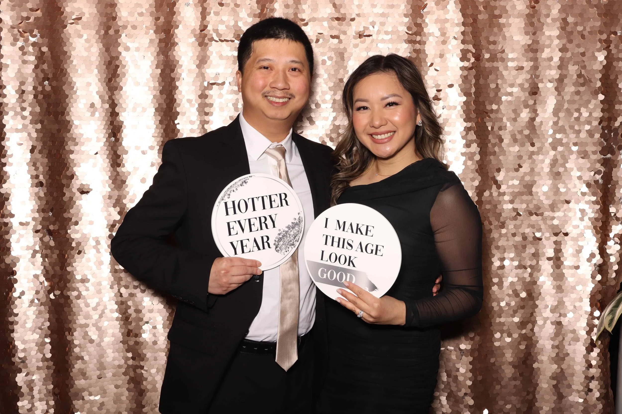 A couple in formal attire holding "Hotter Every Year" and "I Make This Age Look Good" signs at a luxury birthday party photo booth in Seattle featuring a rose gold sequin backdrop.