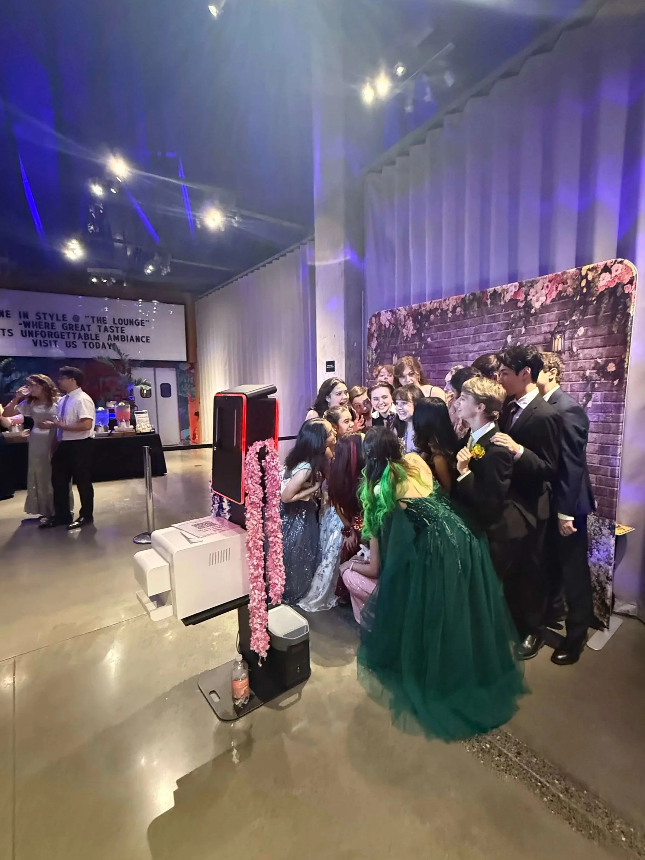 A group of young people dressed in formal attire, some in tuxedos and some in formal dresses, gathered together for a group photo in front of a floral-themed photo booth at a party or event.