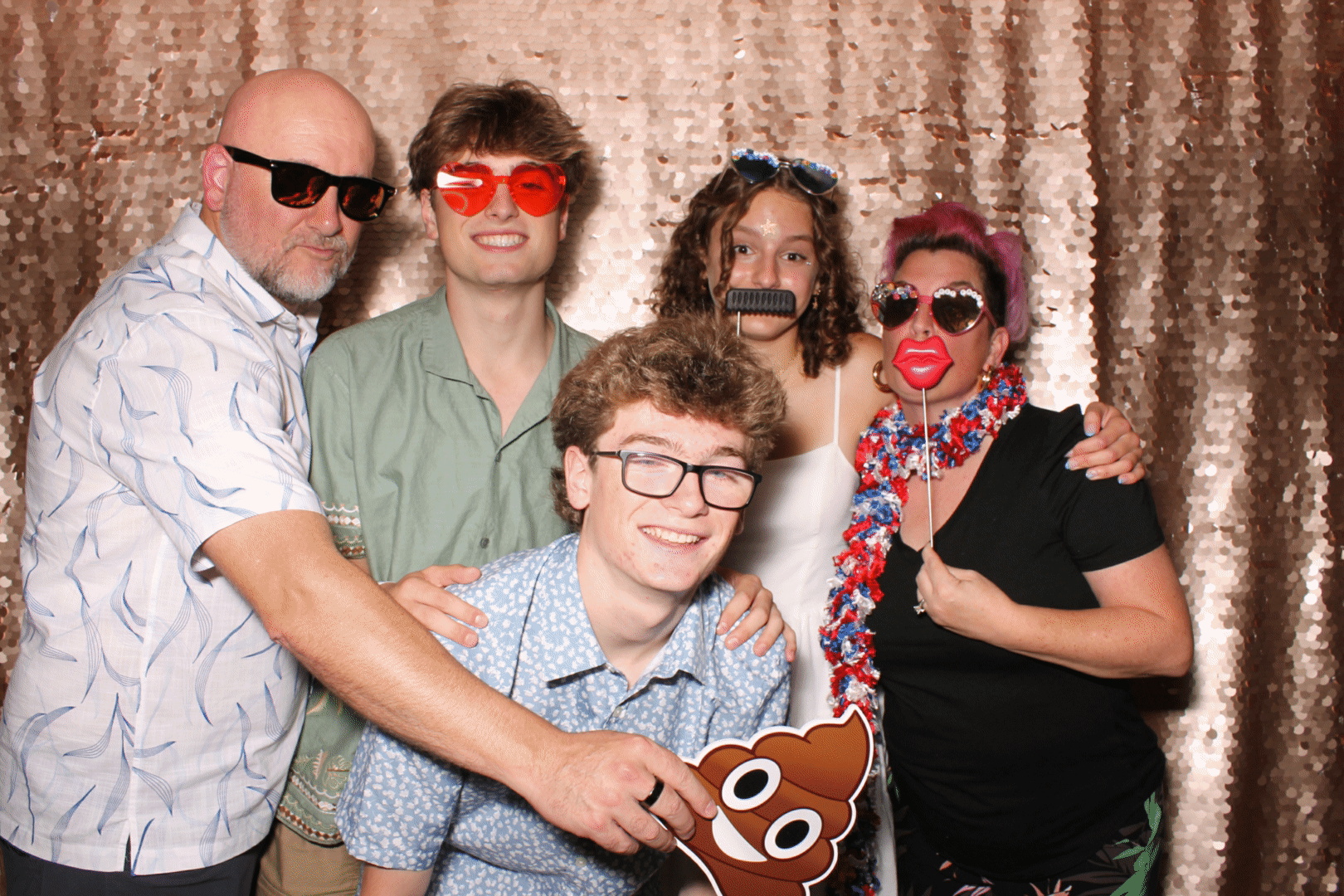 Group of five people at a photo booth, wearing fun glasses and masks, with a shiny, sequin gold backdrop.