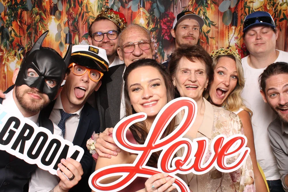Group of people at a celebration, some wearing crowns and hats, holding signs that say 'Love' and '#Groom', smiling and posing for the photo.