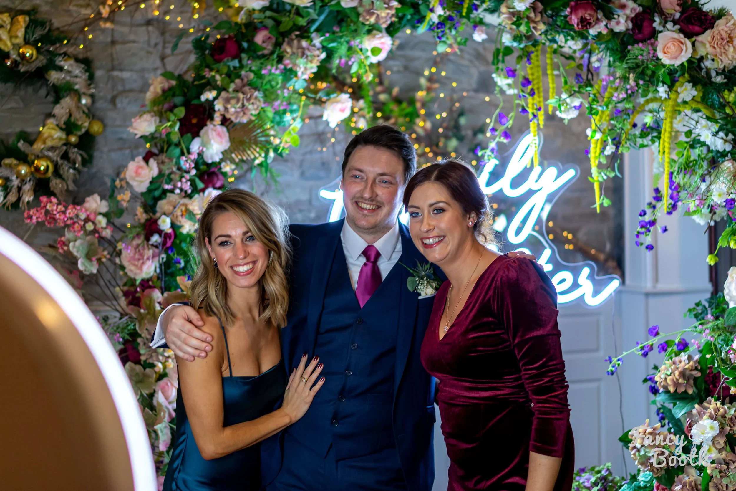 Three guests posing in front of a lush floral arch and neon sign at a luxury wedding photo booth experience by Fancy Booth in Whidbey Island.