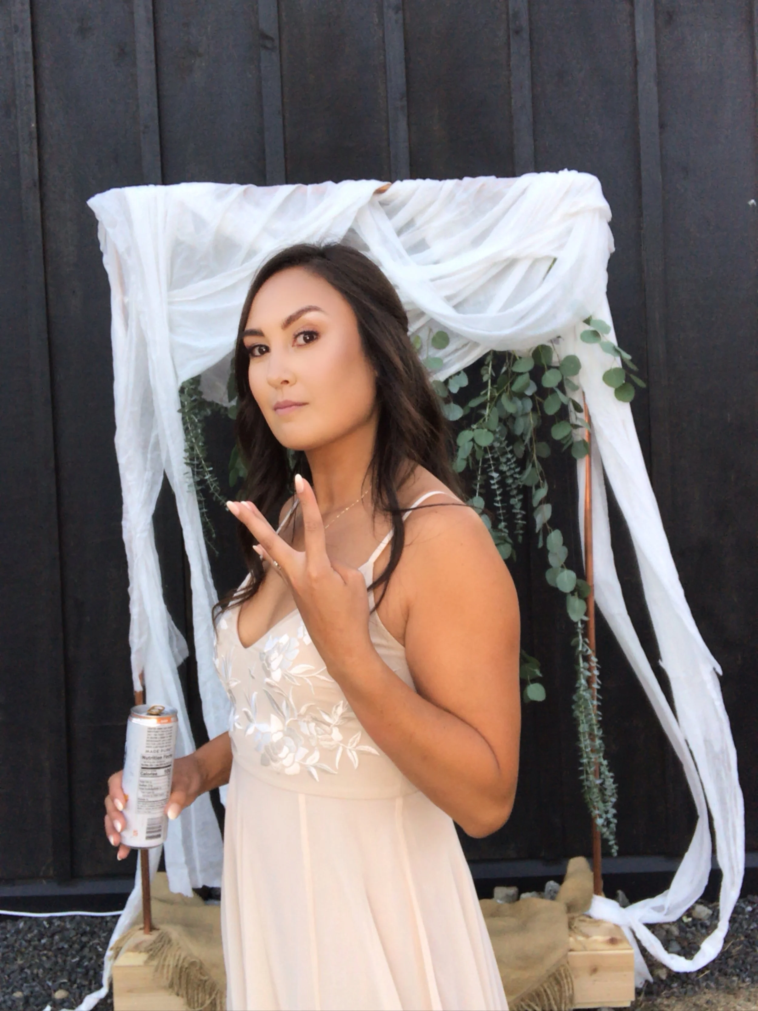 A woman in a cream-colored dress standing outdoors in front of a decorated backdrop with greenery and white fabric.