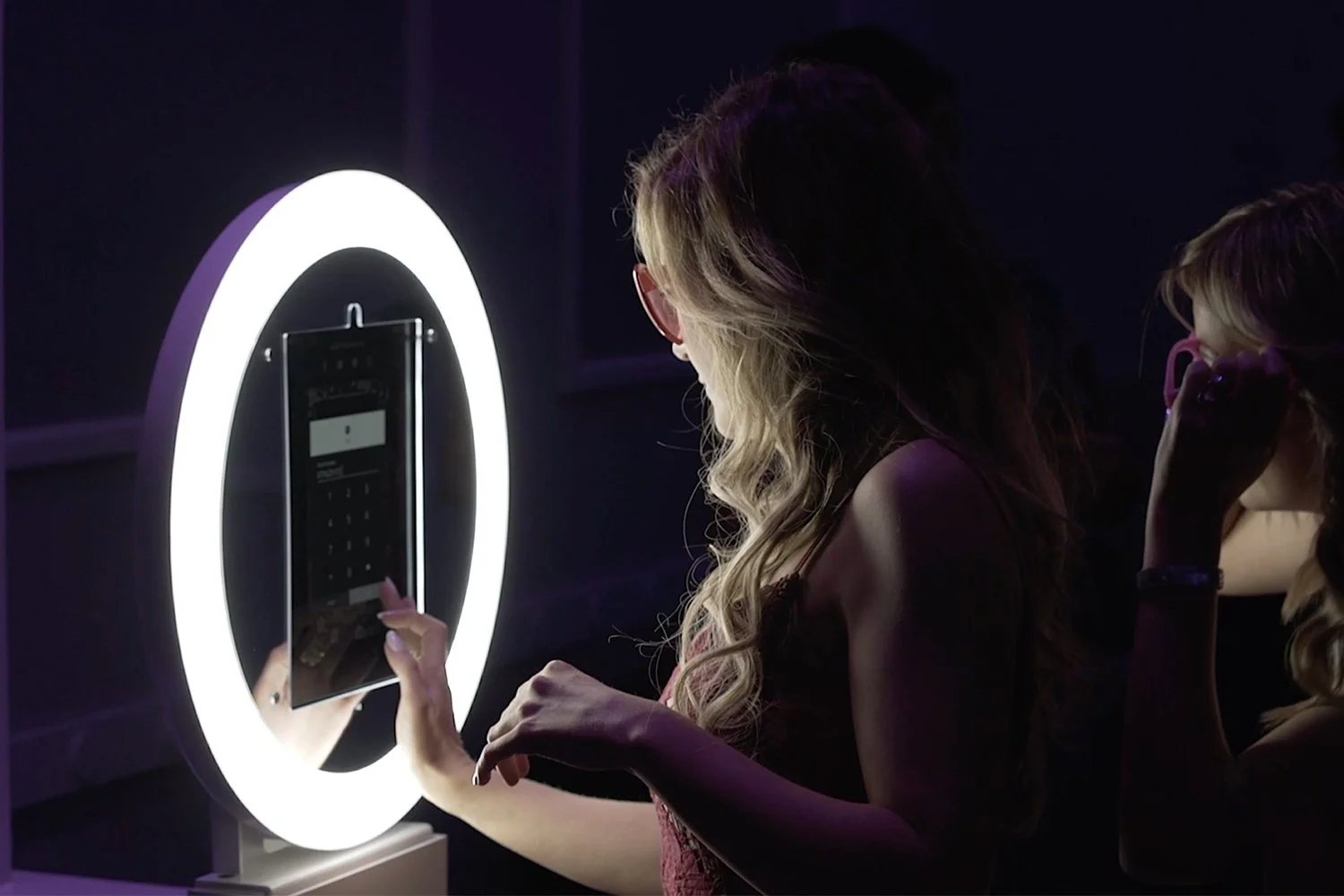 Two women are using a digital photo booth with a circular light frame, taking photos with a tablet in a dark setting.
