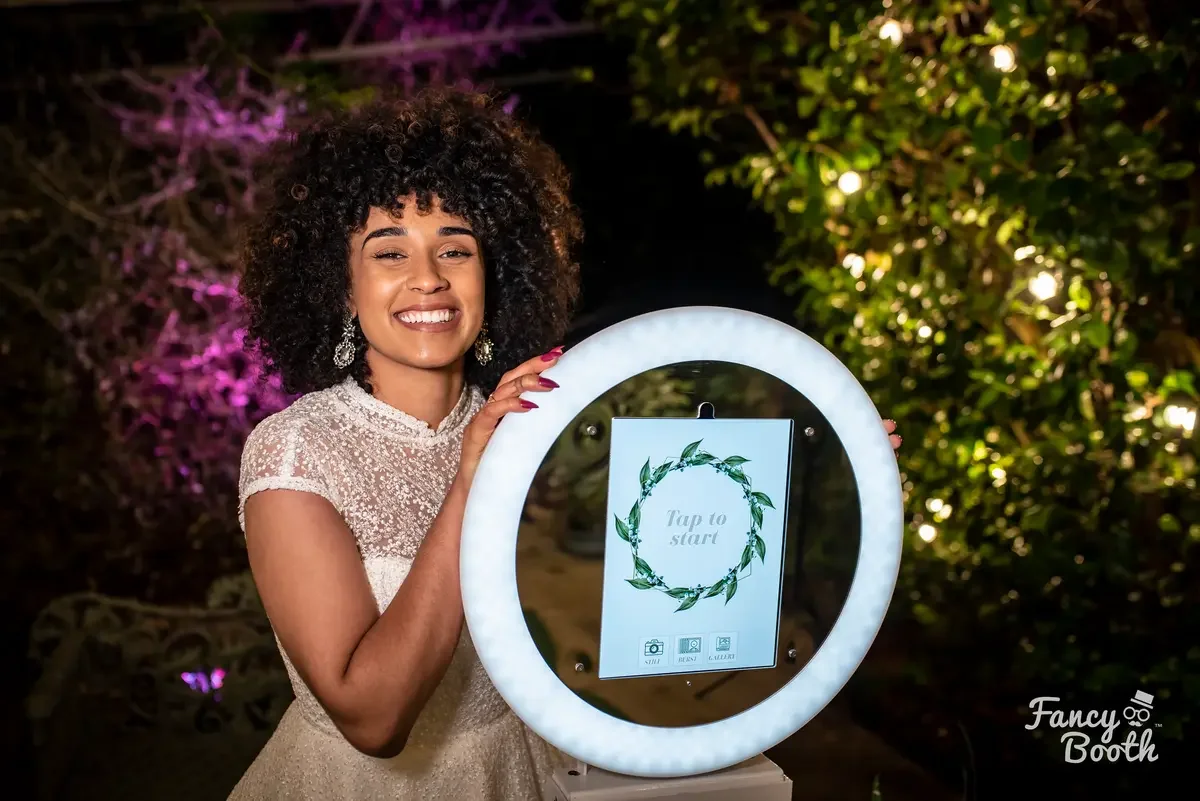 A smiling guest using the Aura digital selfie station at an evening event in Seattle, highlighting the flattering ring light and custom 'Tap to Start' floral interface by Fancy Booths.