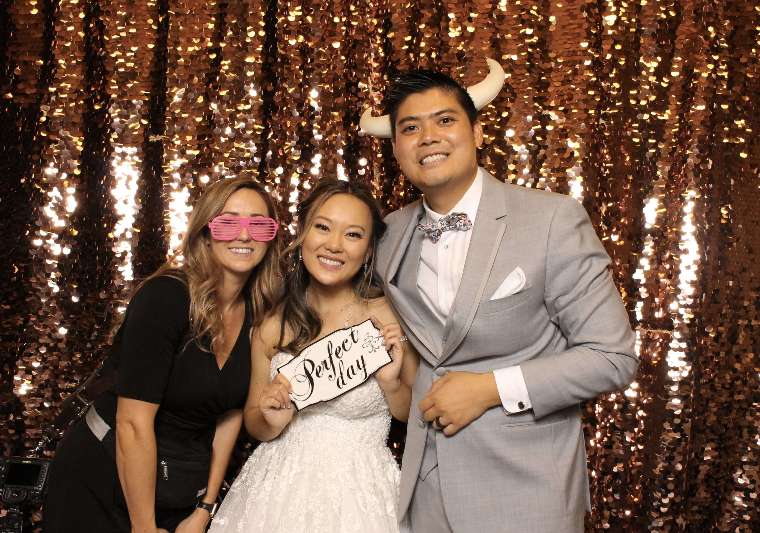Three people at a celebration, with a gold sequin backdrop. The woman in the center is wearing a white dress, holding a sign that says "Perfect day." The man on the right is wearing a light gray suit, a bow tie, and novelty horns on his head. The woman on the left is wearing black and pink shutter shades.