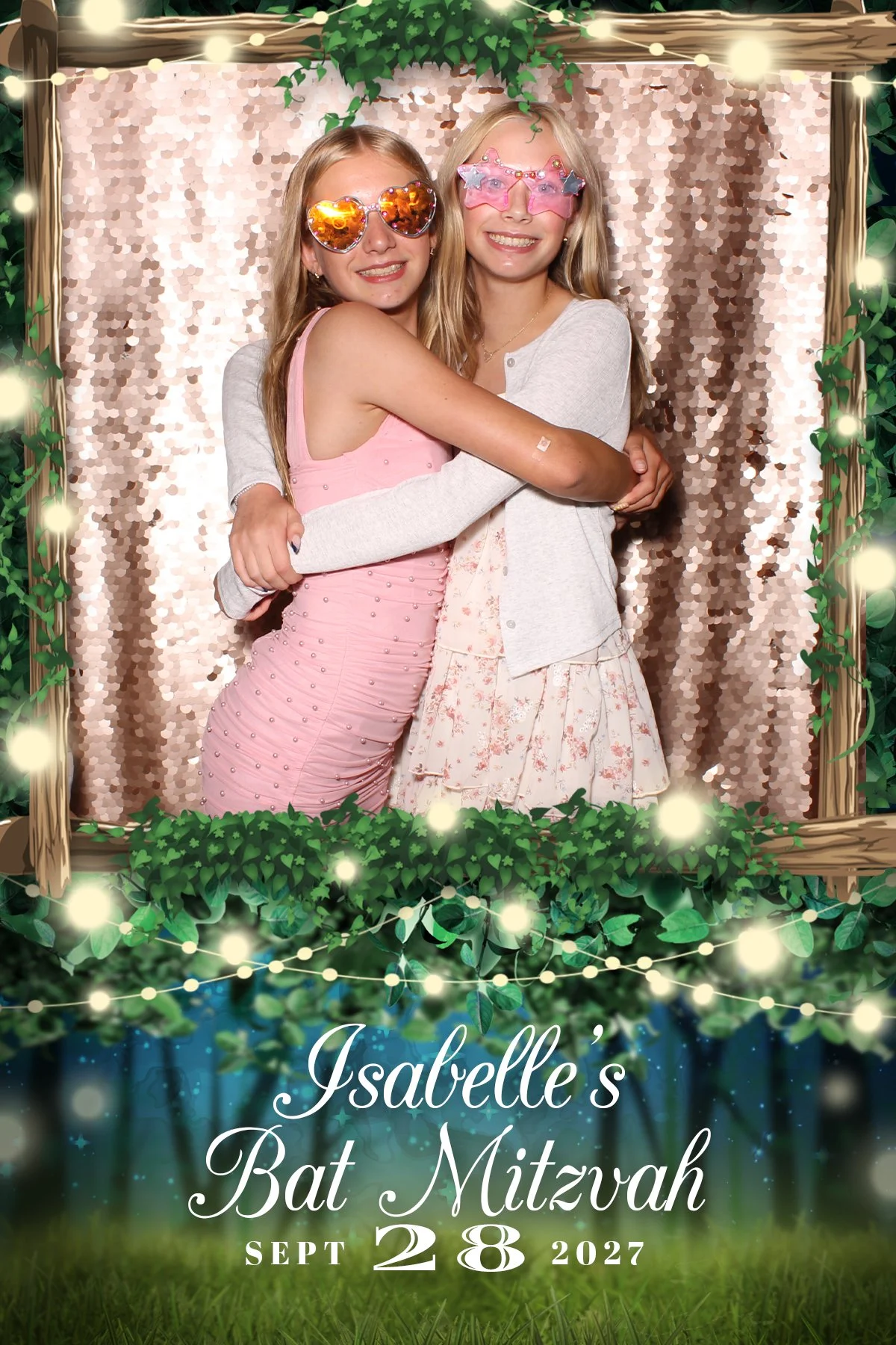 Two girls hugging, one wearing heart-shaped sunglasses and a pink dress, the other wearing star-shaped glasses and a floral dress, in front of a sequined backdrop with a decorative frame and greenery at an event sign reading 'Isabelle's Bat Mitzvah, September 28, 2027.'