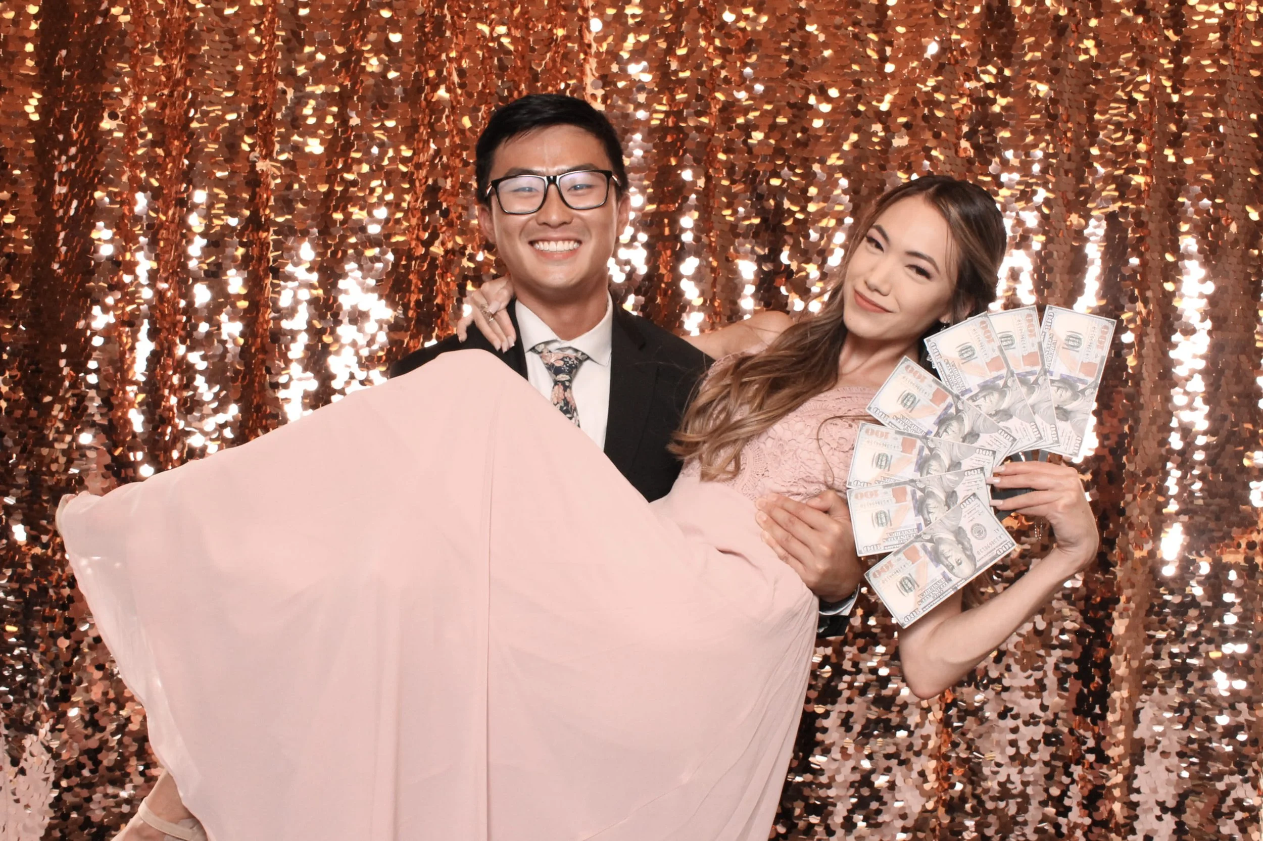 A young couple in formal attire posing with props in front of a rose gold sequin backdrop at a Fancy Booth luxury photo booth in Seattle.