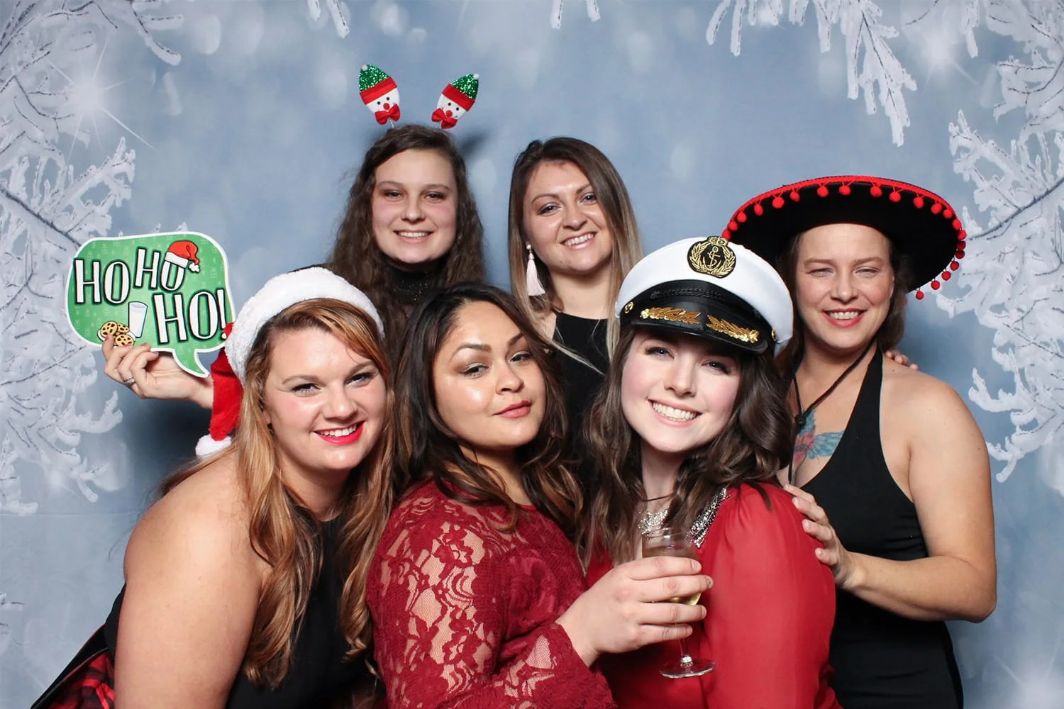 A group of six friends wearing festive hats and holding a "Ho Ho Ho" sign in front of a winter snowflake backdrop at a Fancy Booth holiday party in Seattle.