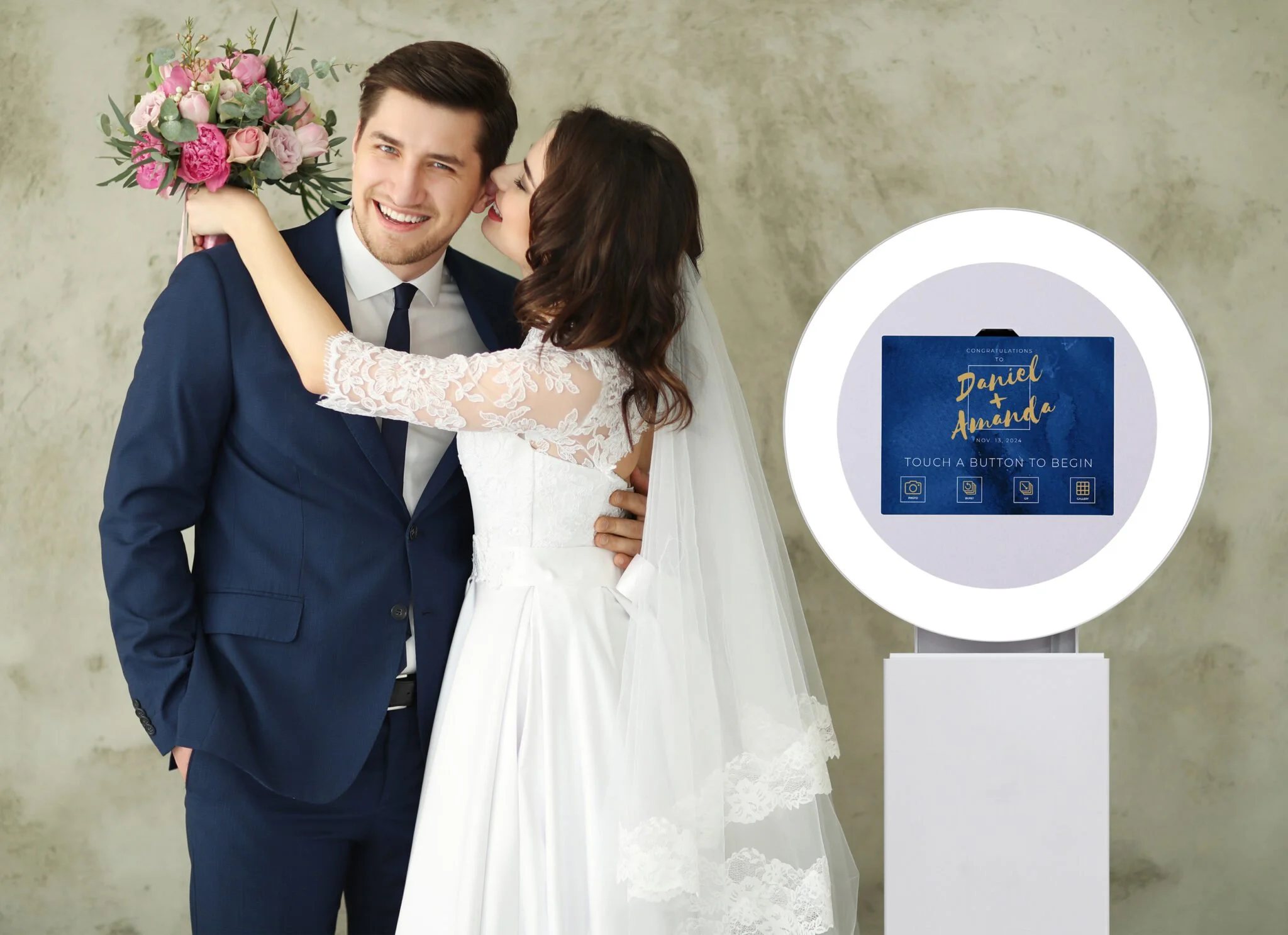 A bride and groom smiling and hugging. The bride, wearing a white wedding dress with lace sleeves and a veil, is holding a bouquet of pink and purple flowers. A device with a digital screen displays a wedding congratulation message for Daniel and Amanda.