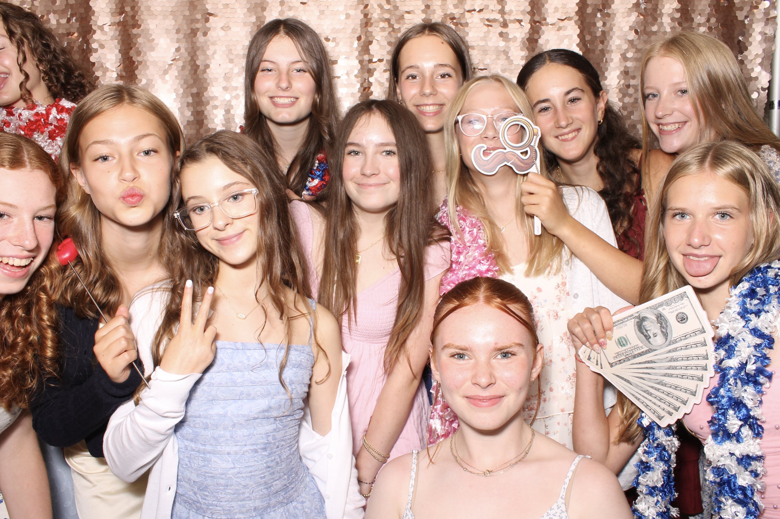 Group of young girls celebrating at a party, some holding props like sunglasses, money, and a lollipop, with a sparkly backdrop.