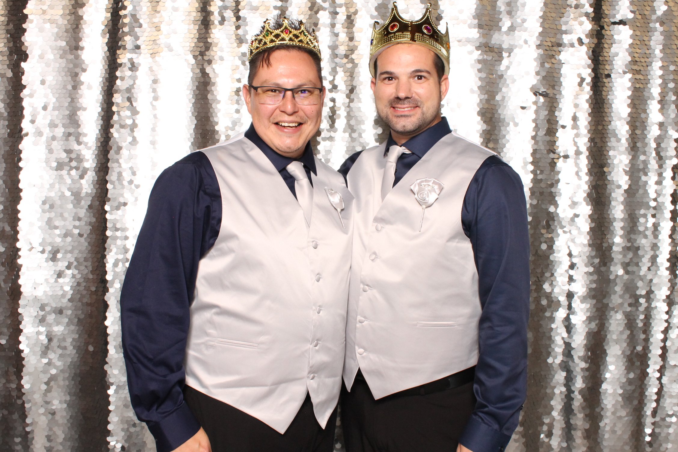 Two men wearing crowns and tuxedo vests over dark shirts, smiling, standing in front of a silver sequin backdrop.