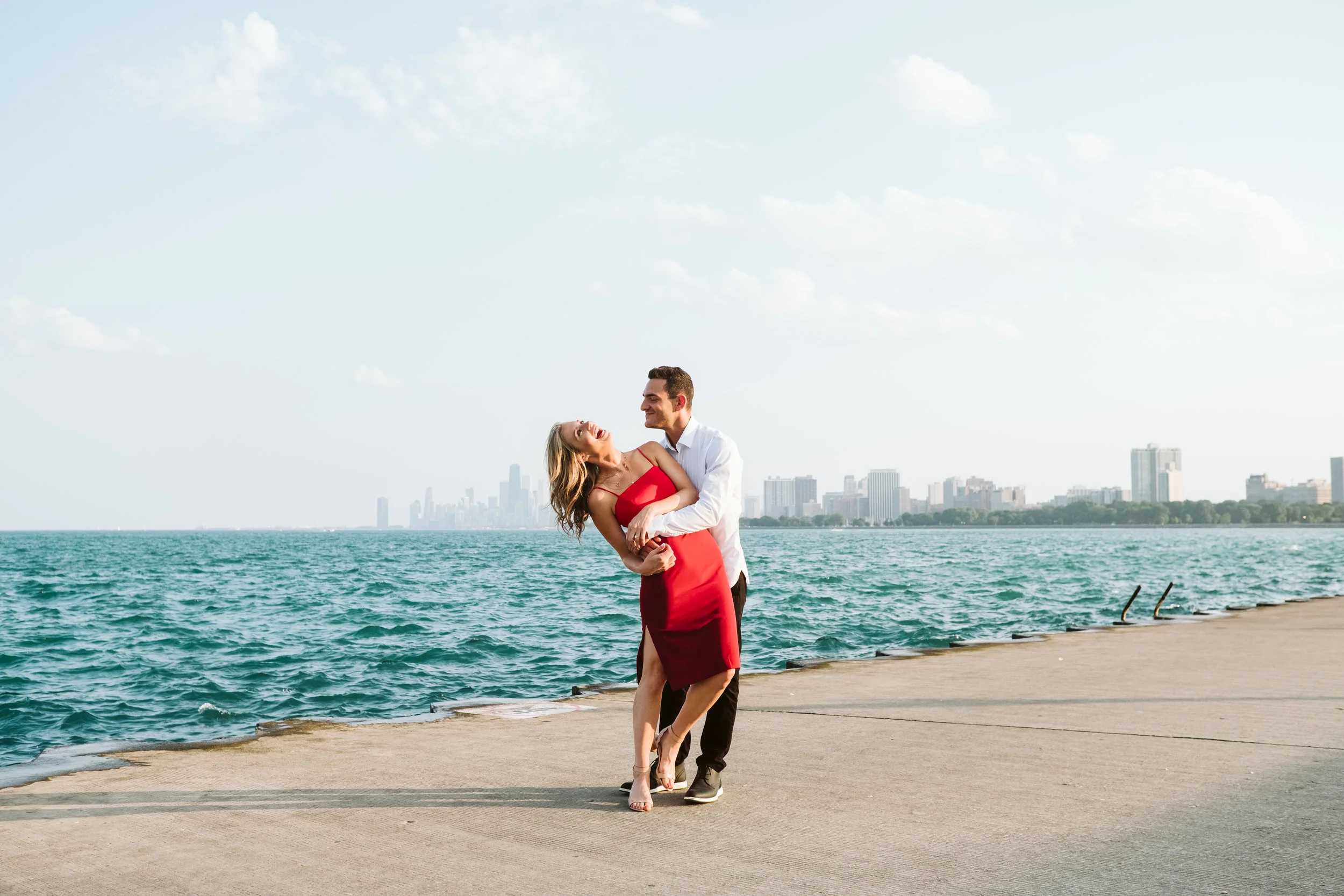 Lake Michigan Engagement Photos featuring a wide photo of the couple. The groom is dipping his soon-to-be bride while they stand on the pier overlooking the Chicago skyline. By Michael & Kristin Chicago Photographer and Videographer 