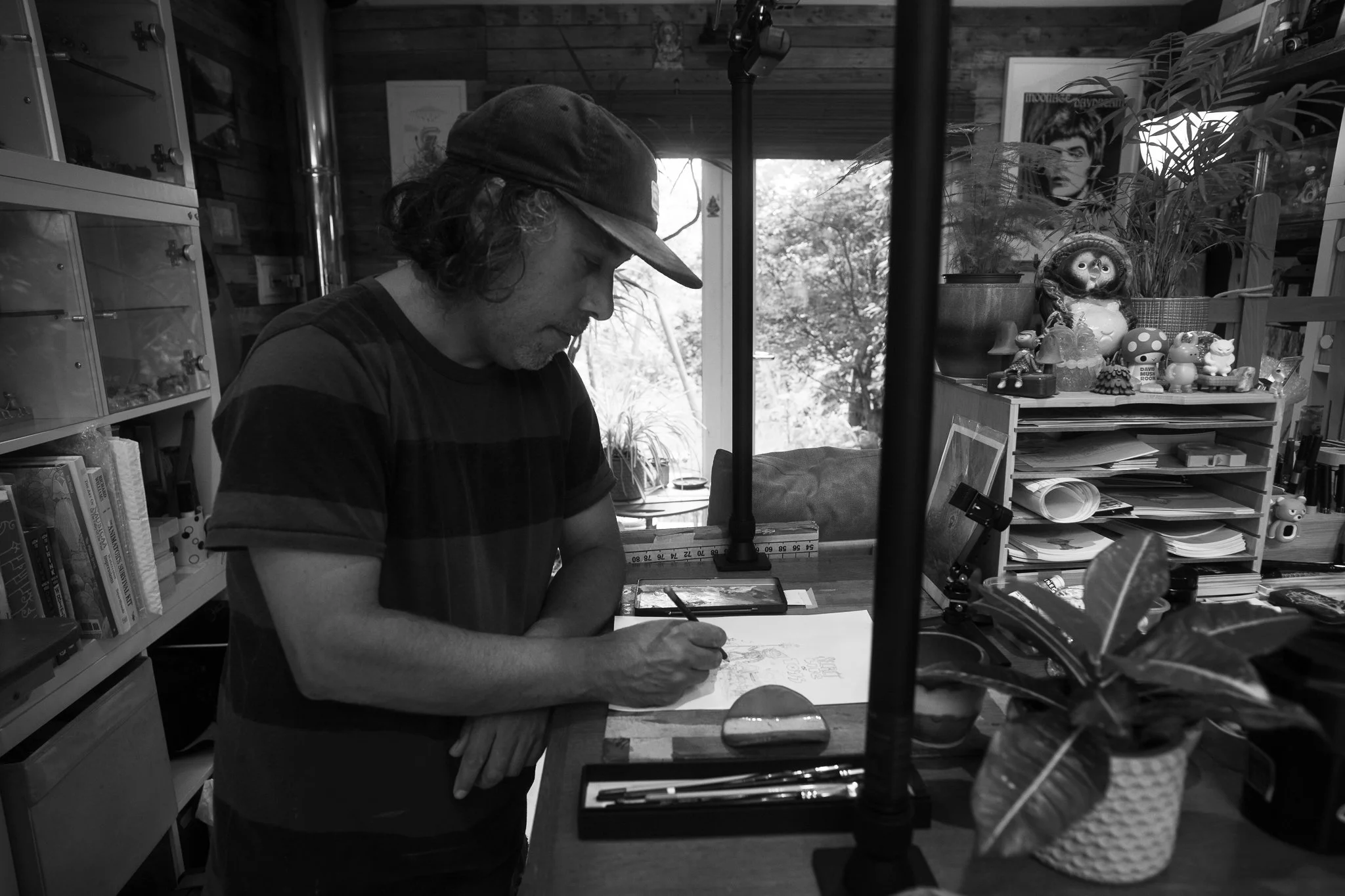 A man with long hair and a baseball cap drawing on a piece of paper at a cluttered desk in a cozy room with plants, books, and decorative items.
