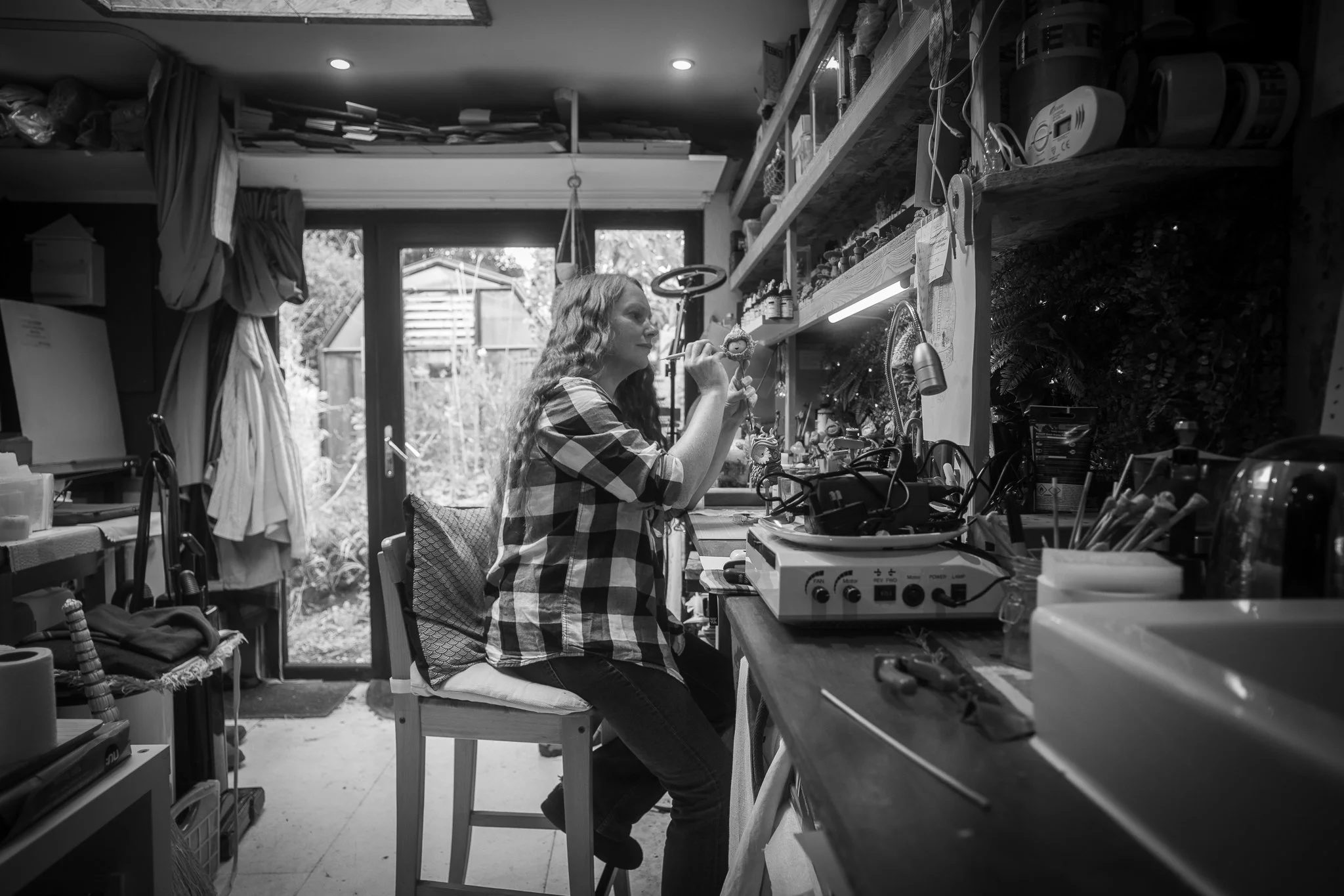 A woman with long wavy hair sitting on a chair in a cluttered workshop or craft room, holding a small figurine close to her face and looking at it. The room has shelves filled with bottles and tools, a window and a glass door revealing a garden outside.