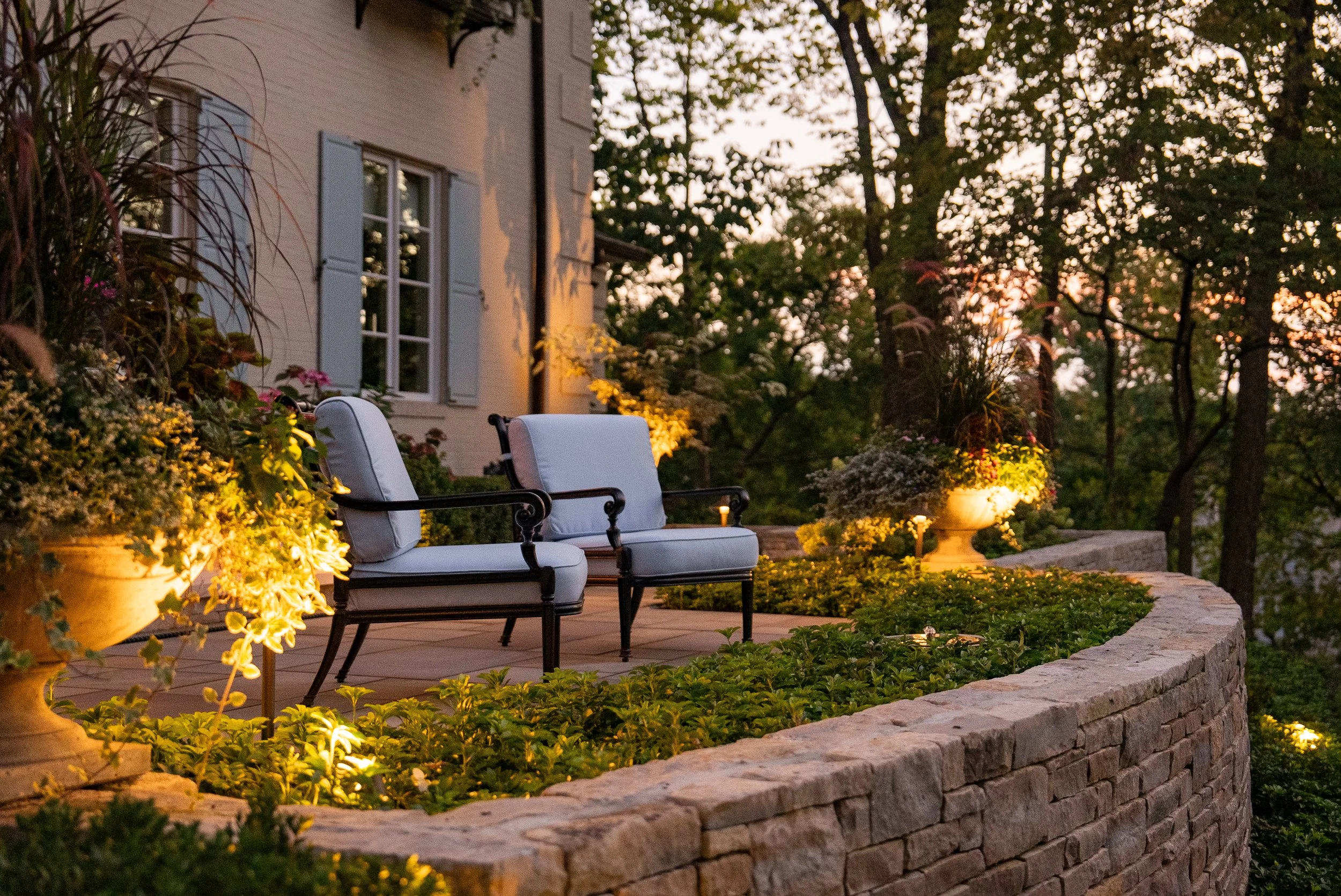 An outdoor patio with two white cushioned chairs, surrounded by potted plants and flowers, illuminated by garden lights at dusk, with a house in the background and trees.