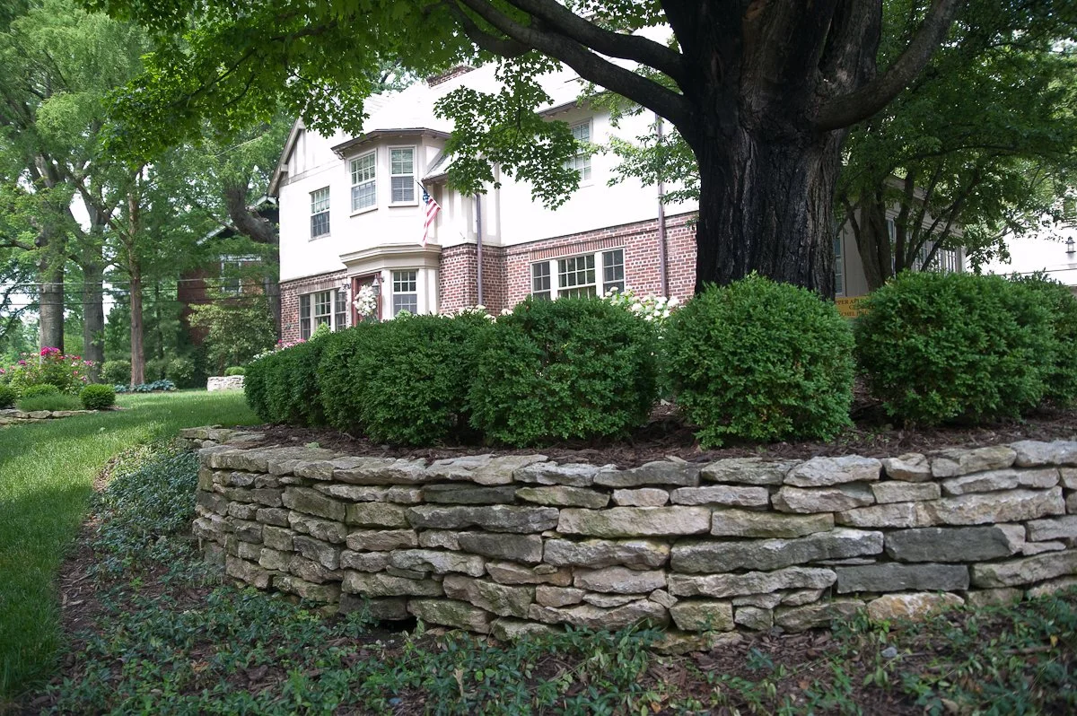 A residential house with a garden featuring a large tree, neatly trimmed bushes, and stone retaining walls.
