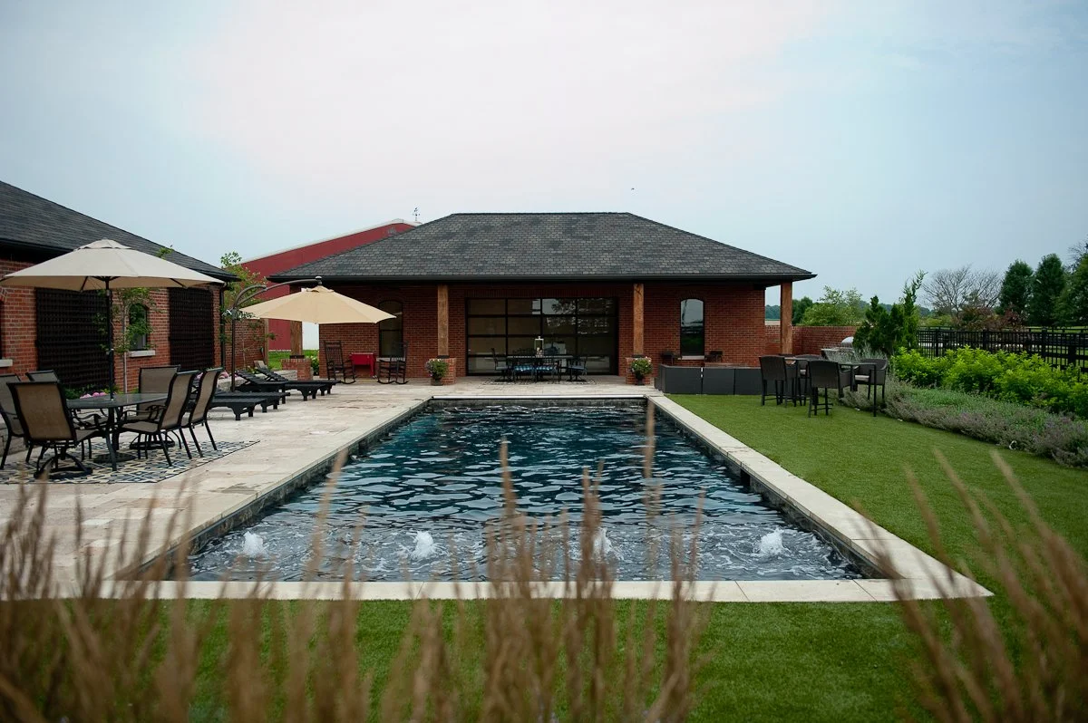 A backyard with a rectangular swimming pool, surrounded by patio furniture, umbrellas, and greenery, with a brick house in the background.