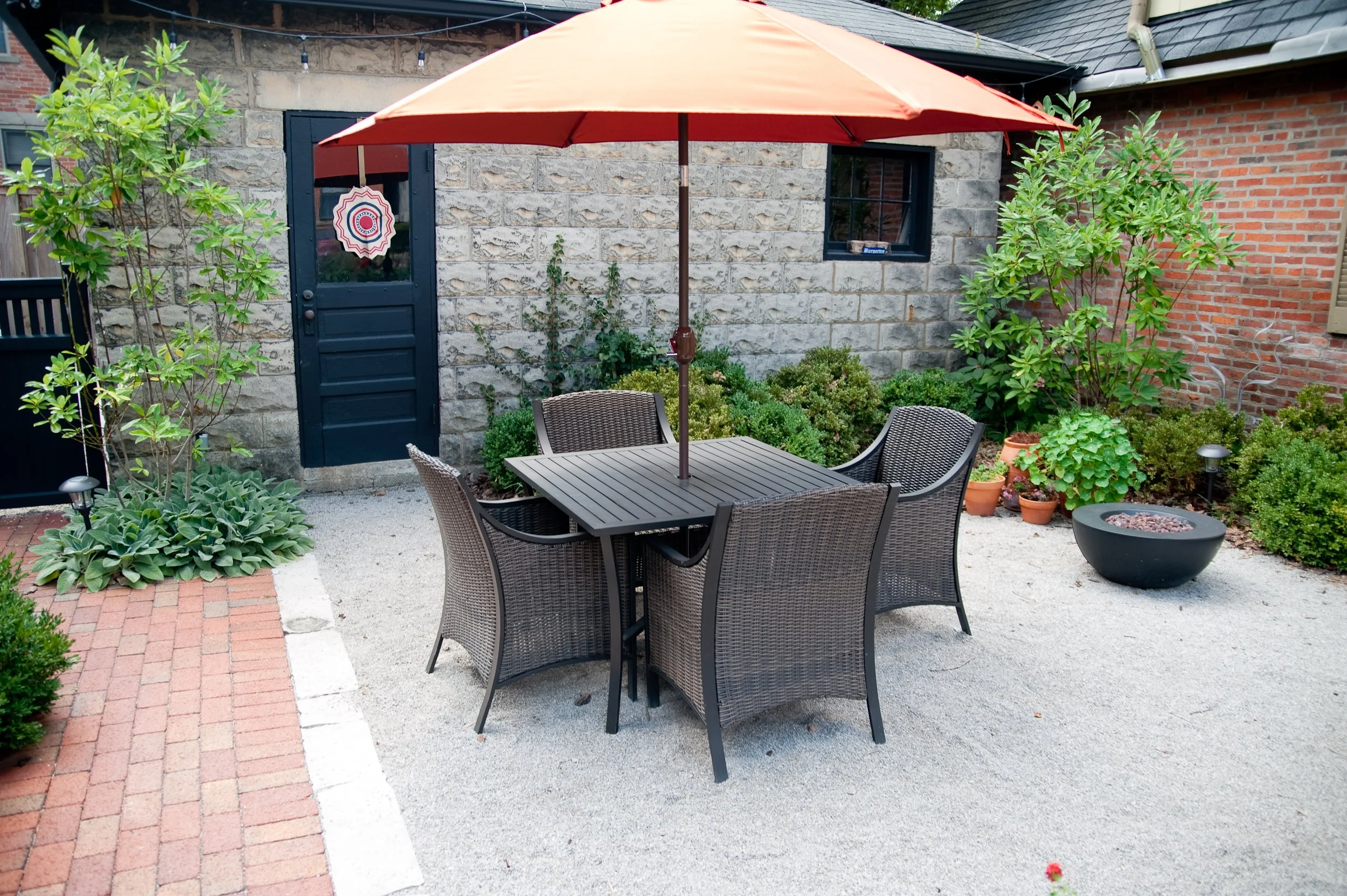 A backyard patio scene featuring a round table with six chairs underneath an orange umbrella. Surrounding the table are various green plants, including bushes and potted plants, with a stone and brick wall in the background.