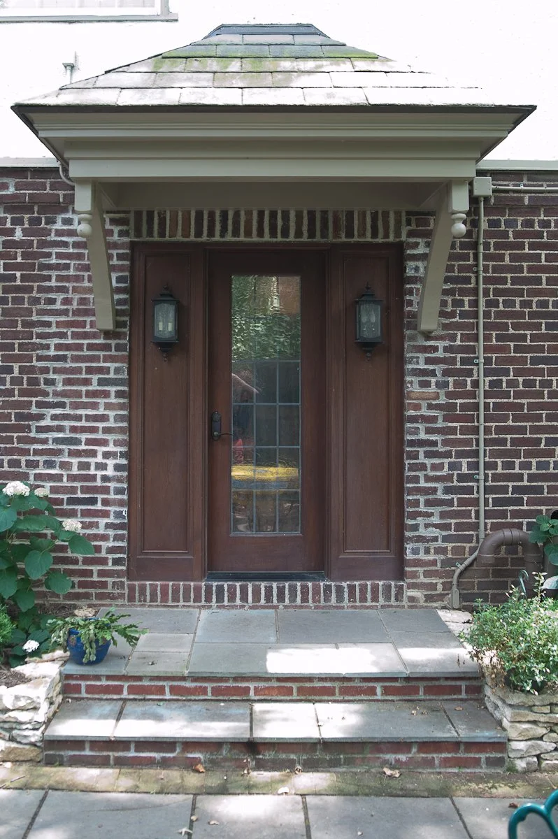 Front entrance of a brick house with a wooden door, two black lantern-style lights, and a small porch with stone steps and plants on each side.