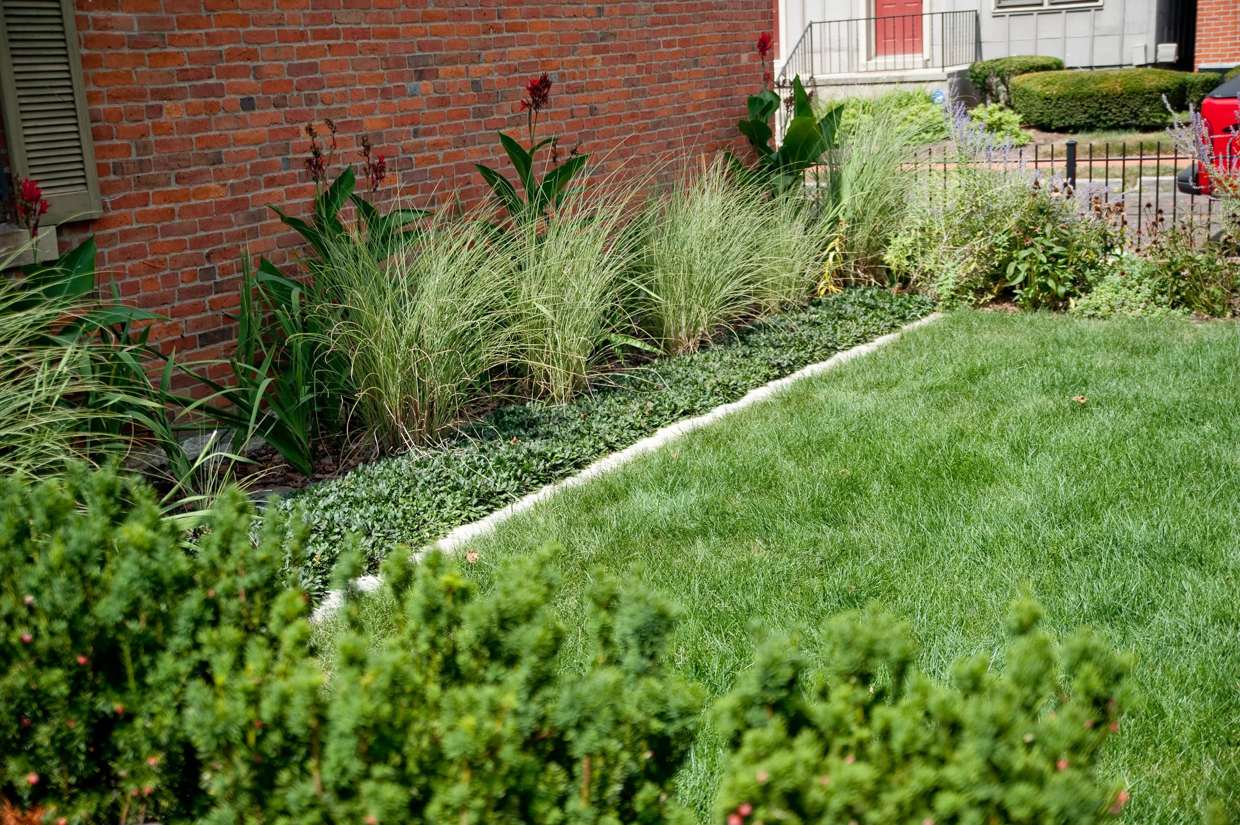 A backyard garden with various plants, a brick wall on the left, a small lawn in the foreground, and a house with a porch and a red vehicle in the background.