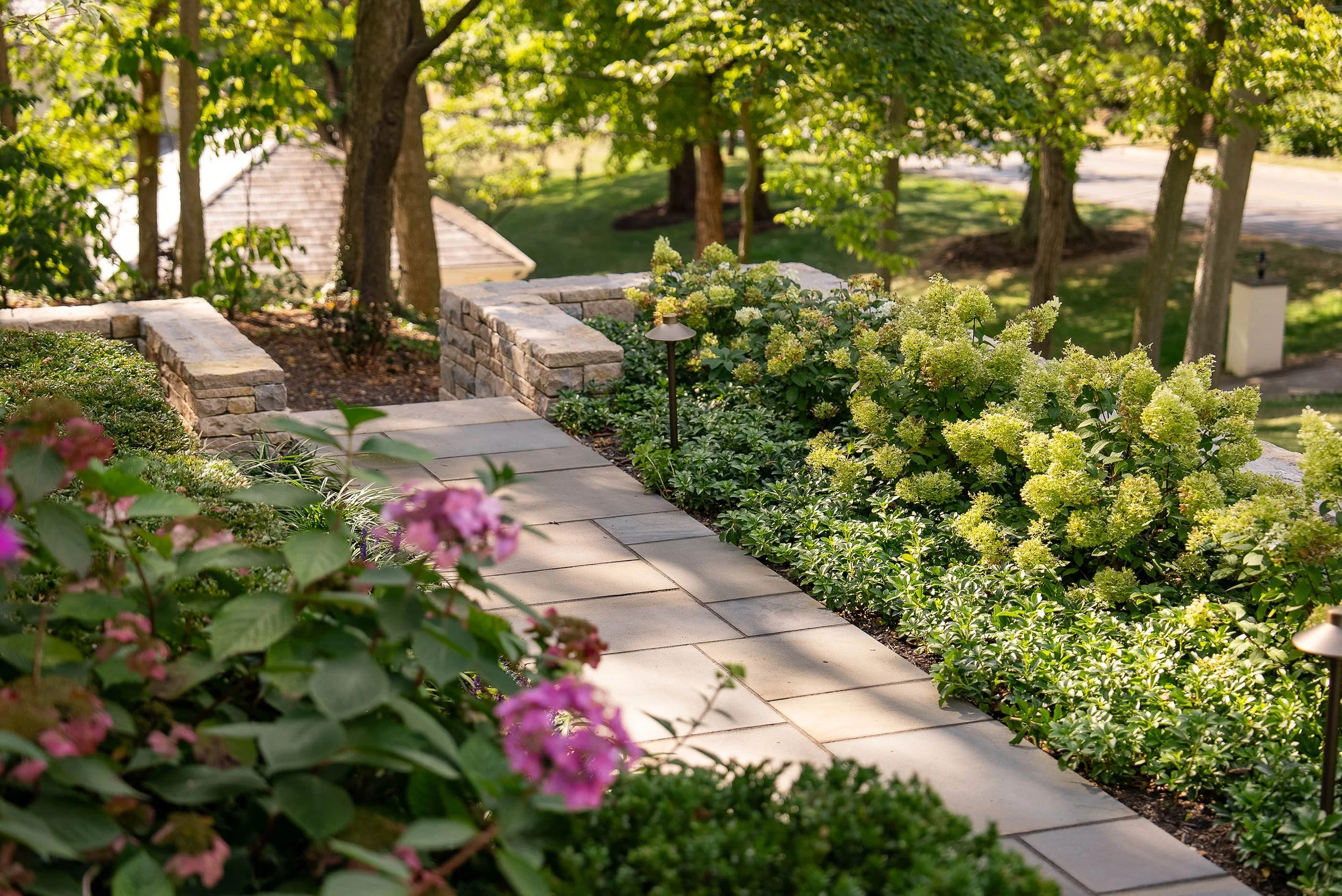 A landscaped garden with a stone pathway, lush green bushes, flowering plants, trees, and a small lamp post.