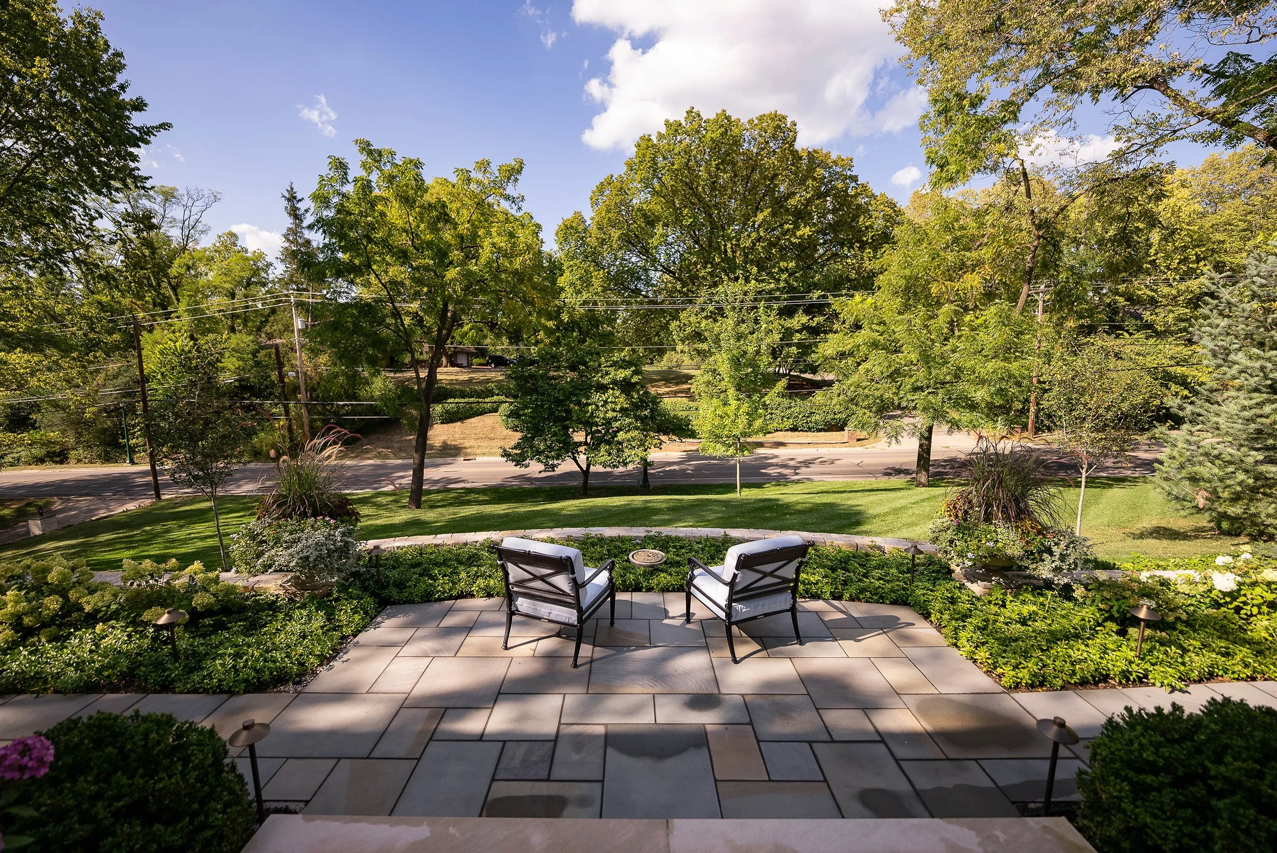 View from a patio overlooking a green yard with trees and road beyond, two chairs and small table on the patio.