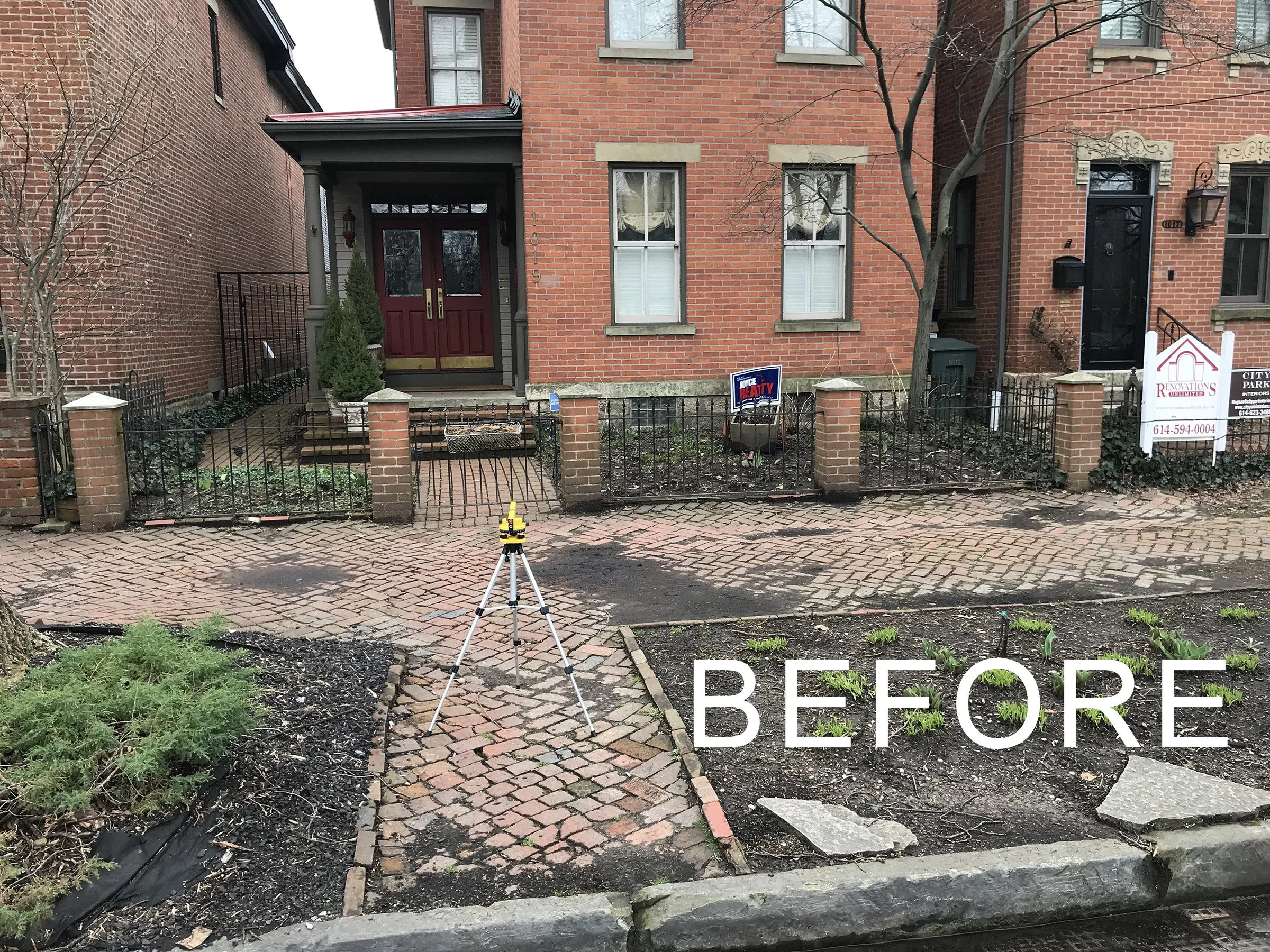 Front of a brick house with a small garden in the yard, a warning sign for real estate, and a tripod surveying instrument on the sidewalk. The house has a dark red door and multiple windows, and the yard appears to be in the process of landscaping or renovation.
