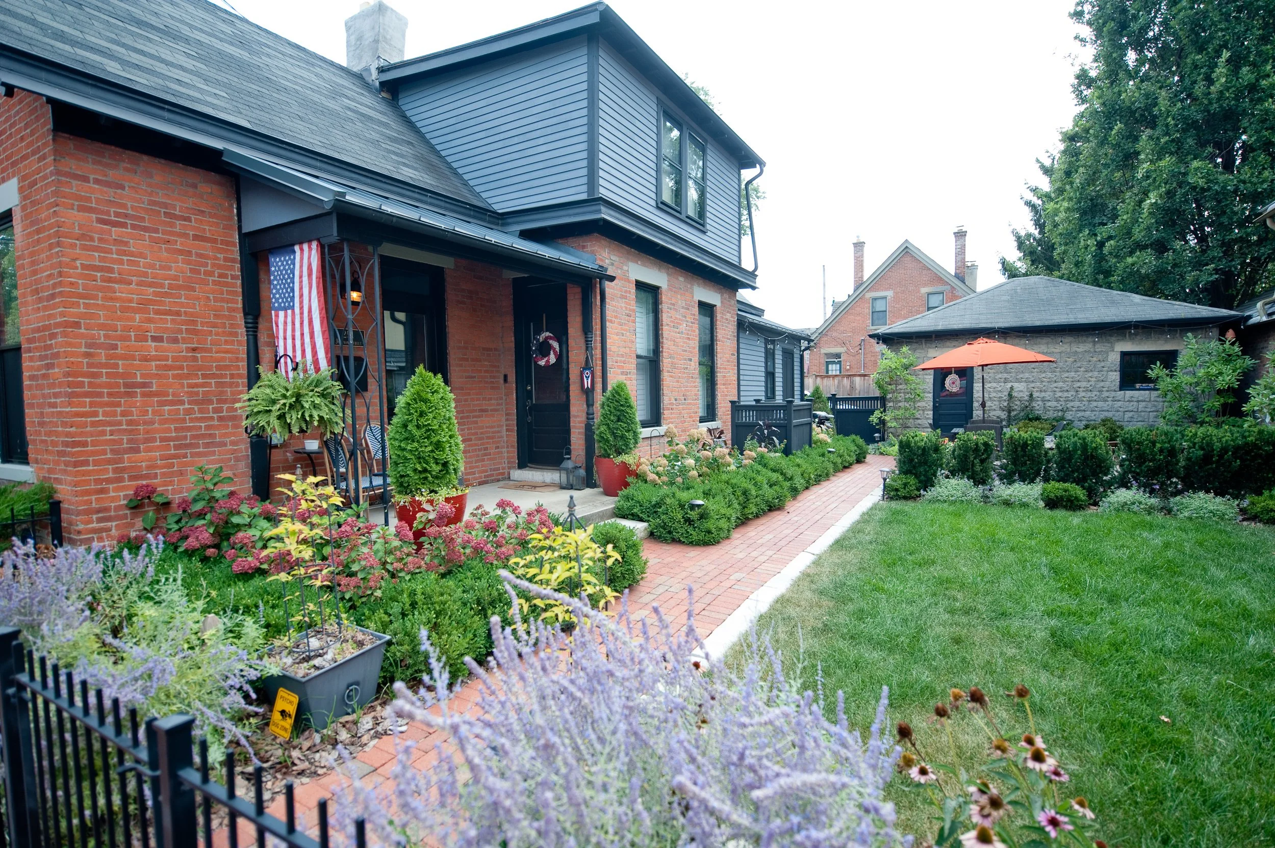 A backyard with a brick and blue house, a brick pathway, lush green grass, and flower beds with purple and pink flowers; a large umbrella provides shade near a small building in the yard.