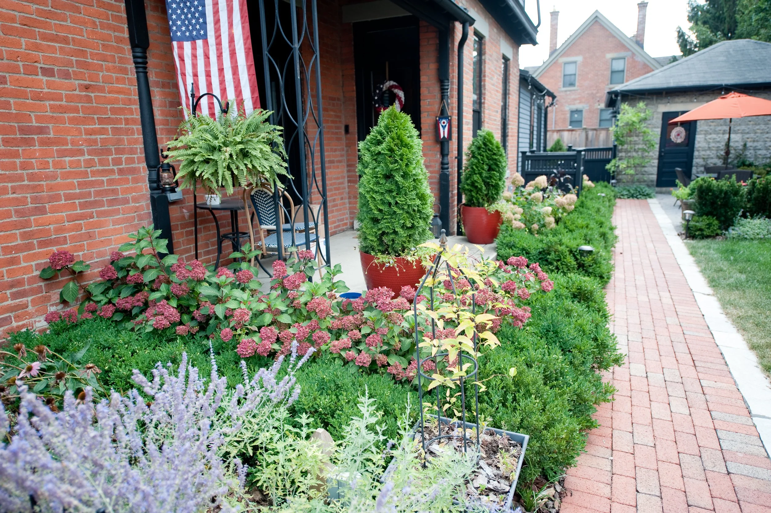 Front yard garden with flowering plants, potted shrubs, and a brick sidewalk leading to a house decorated with an American flag and patriotic decorations.